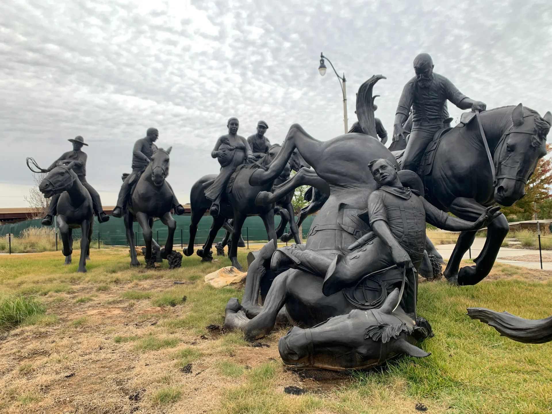 A dynamic black sculpture depicts several riders on horseback in various active poses. Some horses are rearing, and one appears to be mid-fall, with a rider clinging on. The setting is an outdoor grassy area under a cloudy sky. A streetlamp and building are visible in the background.