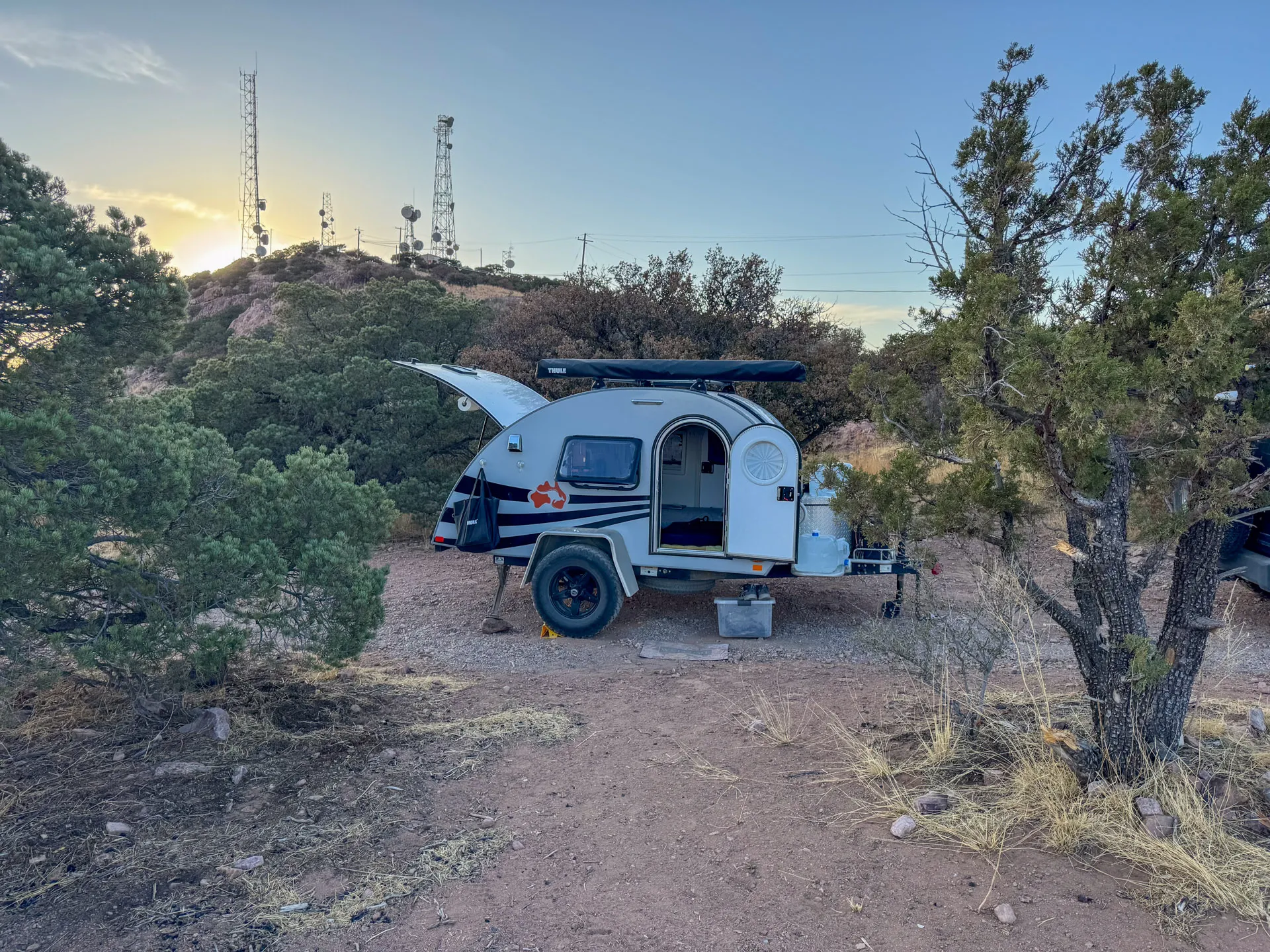 A small teardrop camper trailer is parked at a dirt campsite off North Juniper Flats Road, surrounded by trees and brush. Its back hatch reveals a compact kitchen. Tall towers rise in the background as the Bisbee sunset casts a warm glow across the scene.