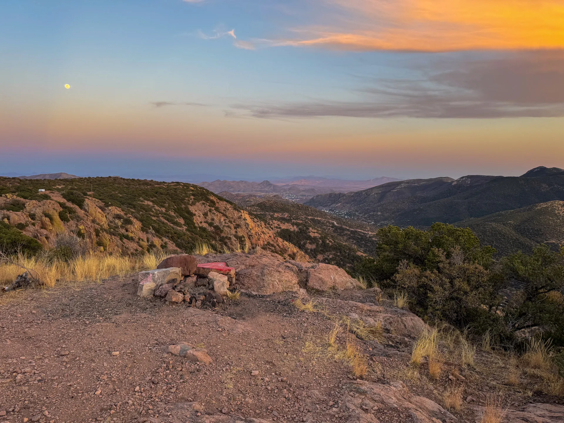 A scenic mountain landscape at sunset near North Juniper Flats Road, with rocky terrain and sparse grass in the foreground. The sky shows blue, purple, and orange hues—and the moon—while distant valleys stretch toward Bisbee under a colorful sky.