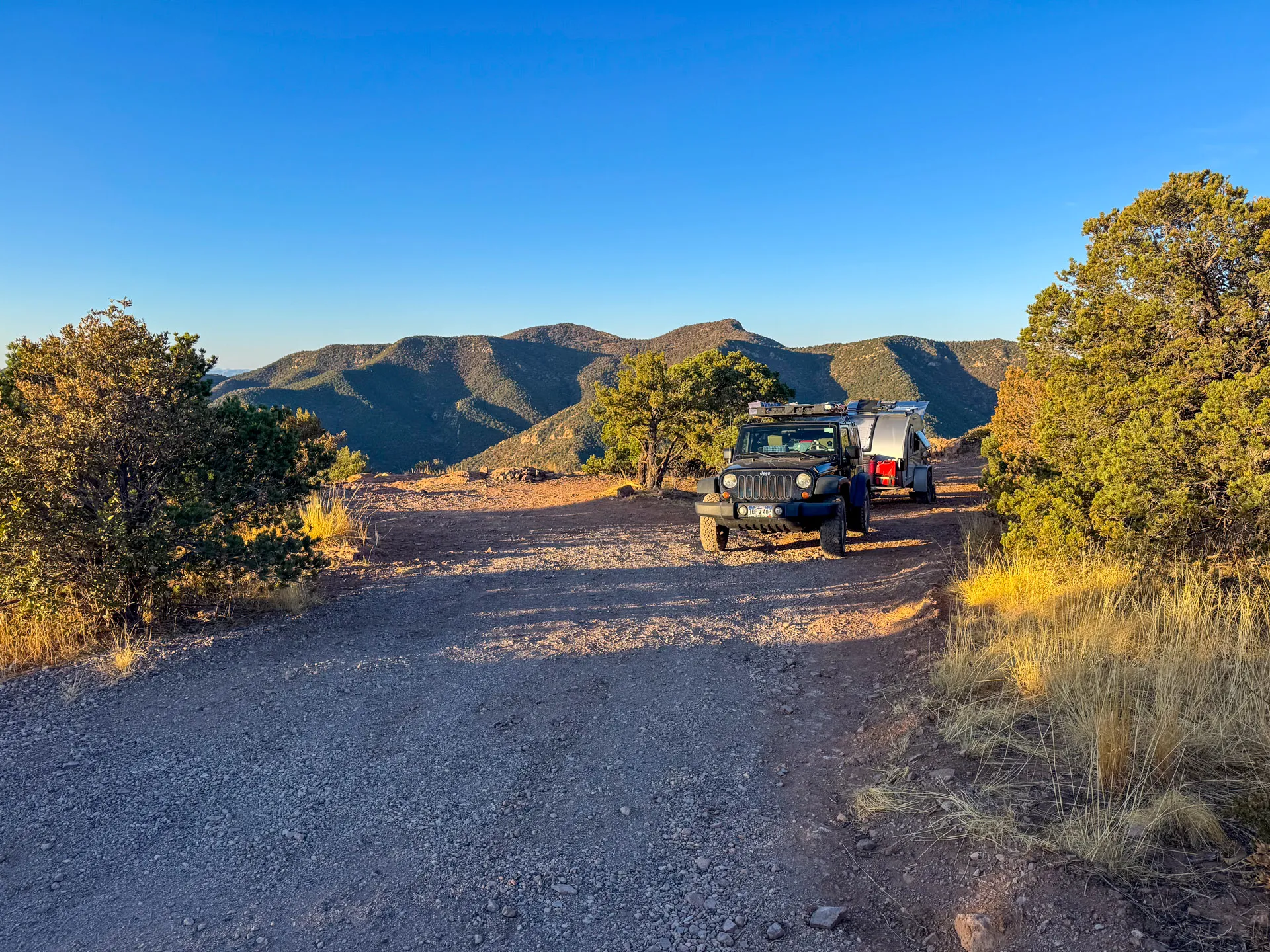A black Jeep SUV towing a small silver camper trailer is parked on a gravel clearing along North Juniper Flats Road. Sunlight casts shadows on the ground, while green mountains rise under a clear blue sky—perfect for camping near Bisbee.