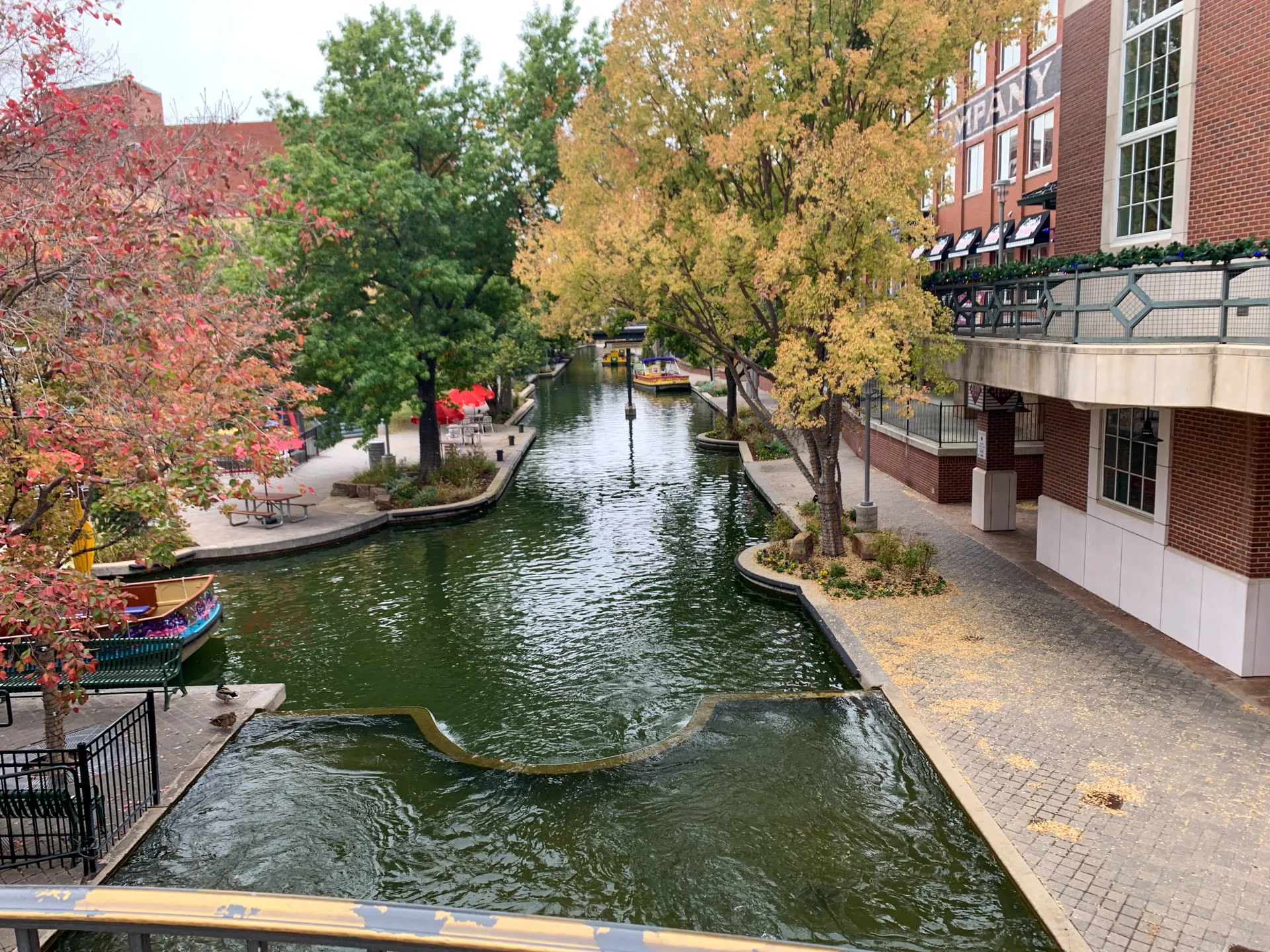 View of a tranquil canal lined with trees in autumn colors, including green and orange foliage. Brick buildings with large windows flank the waterway, and a footbridge crosses overhead. A few small boats are docked along the canal, and leaves are scattered on the paved walkway.