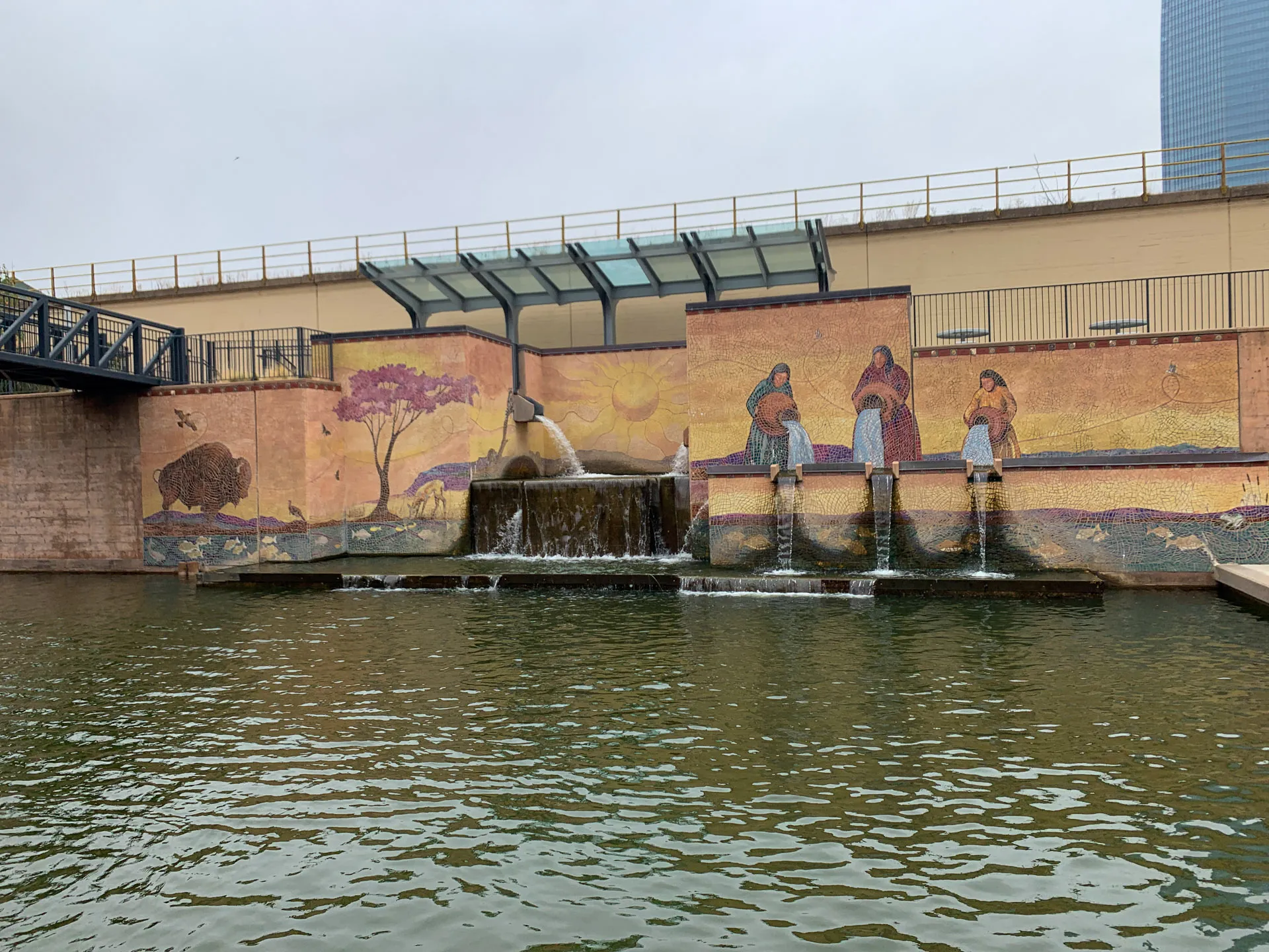A mural with indigenous figures and nature scenes adorns a wall above a cascading waterfall. Four vertical spouts pour water into a pool below. The painting features people, a sun, a tree, and a buffalo. A modern building and railing are visible in the background under a cloudy sky.