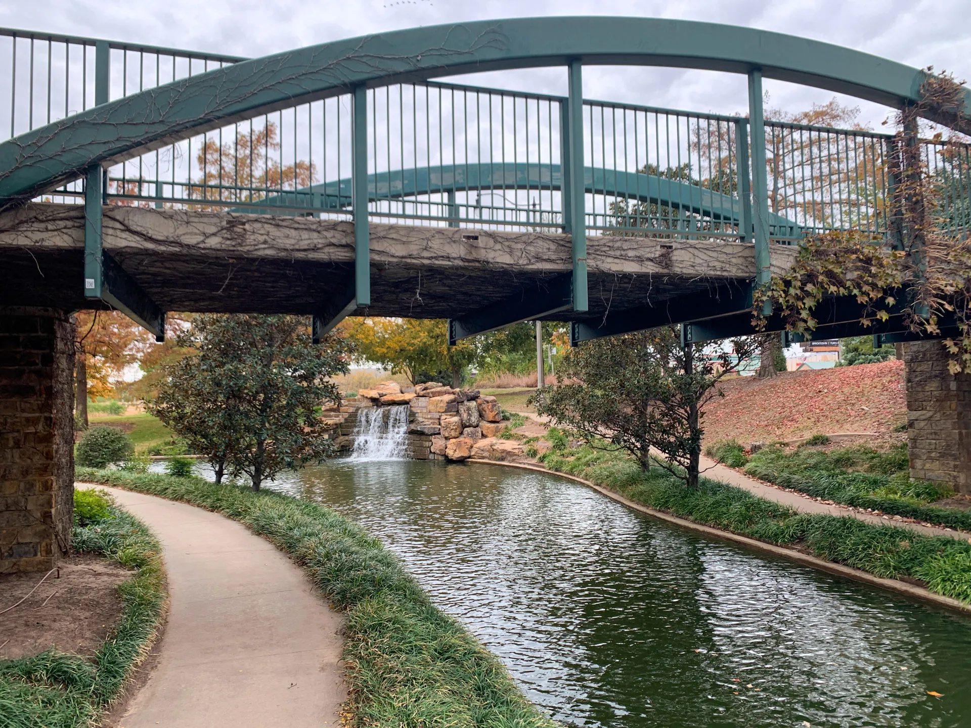 A tranquil scene of a park with a curving path beside a narrow, green river. A green metal bridge arches over the river, with a small waterfall and rocks in the background. Trees with autumn foliage and greenery line the riverbanks under a cloudy sky.