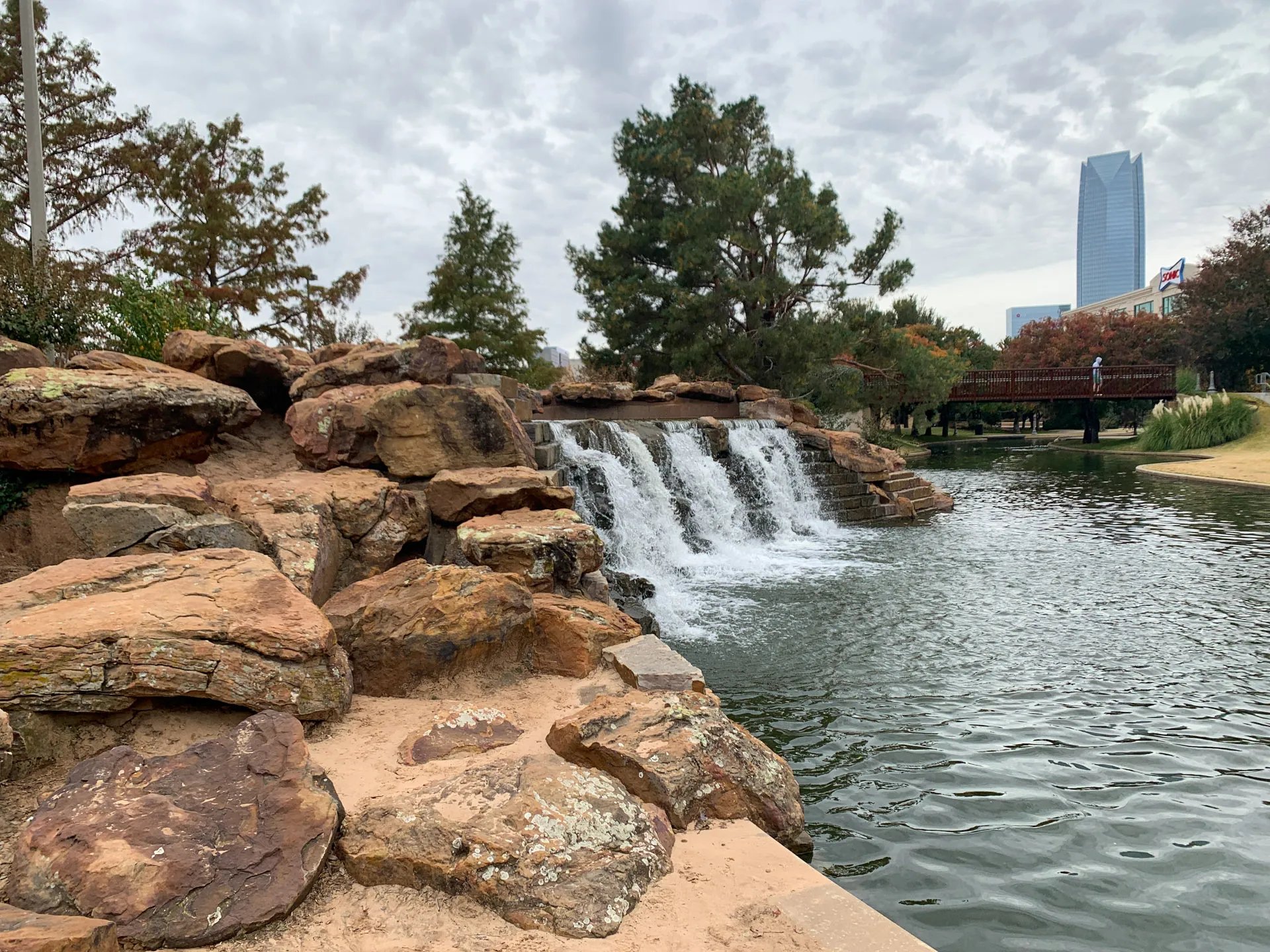 A tranquil park scene with a cascading waterfall over rocky ledges into a pond. Trees with autumn foliage surround the water. A wooden bridge in the background spans the pond, with a tall building visible under a cloudy sky. A person stands on the bridge, observing the view.