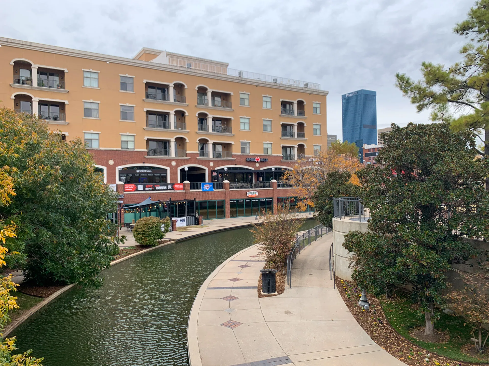 View of a canal with a concrete walkway on the right and a four-story brick and stucco building on the left. The building has balconies and retail spaces. Trees with green and orange leaves line the path. A tall modern building stands in the background under a cloudy sky.