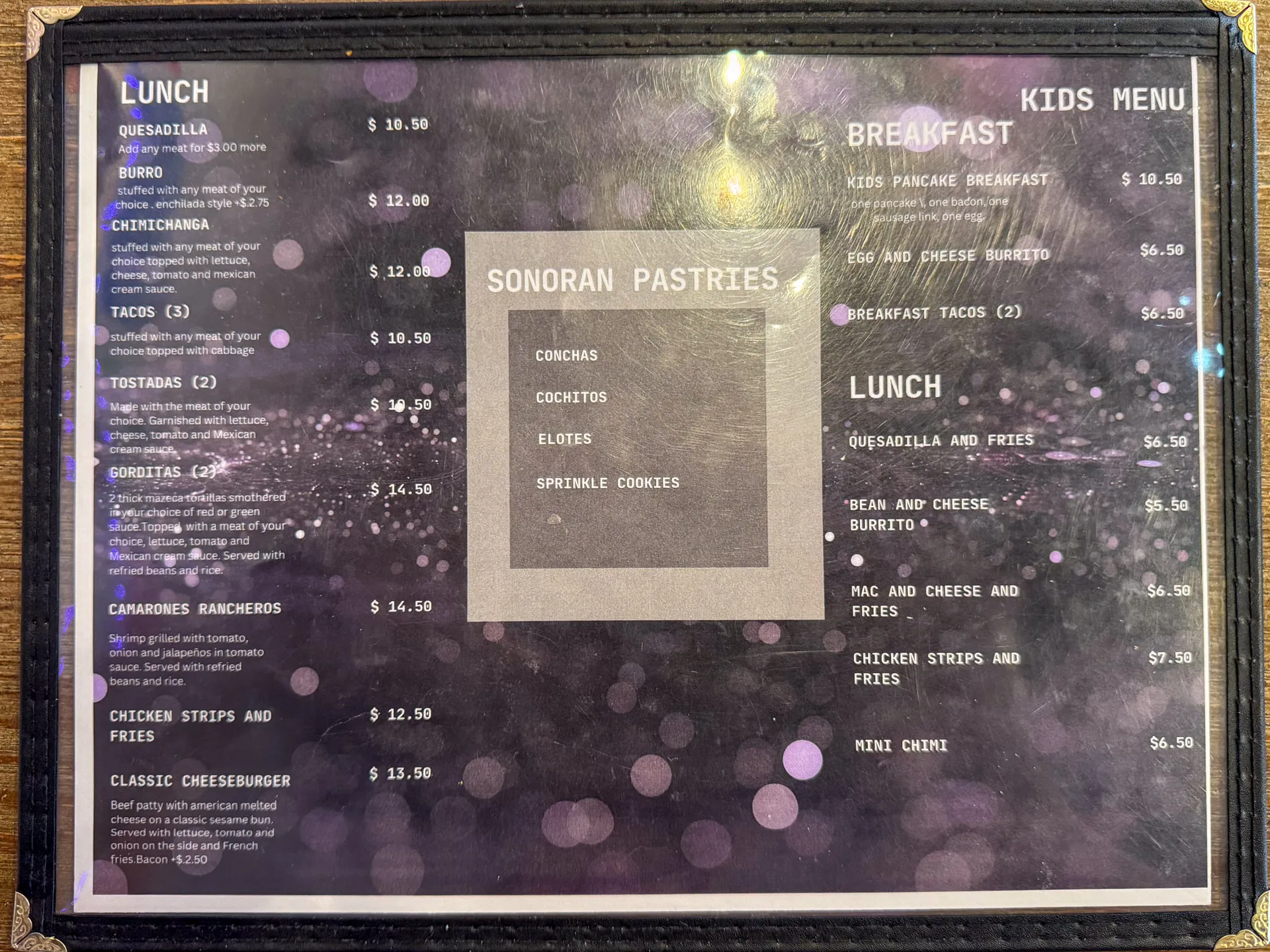A menu with a purple, bokeh background lists food items in categories: Lunch, Sonoran Pastries, Breakfast, and Kids Menu. Prices are in dollars. Items include quesadillas, tacos, tostaditas, chimichangas, and pastries like sprinkle cookies.