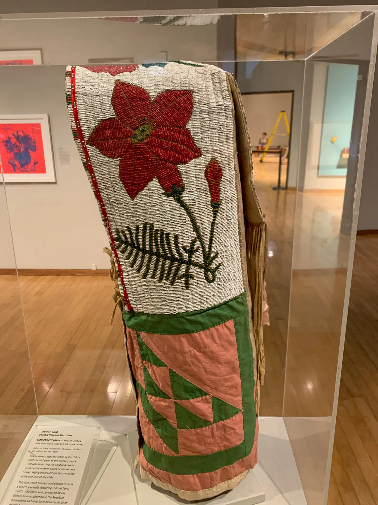 A vibrantly colored cradleboard featuring a large red flower design. The board is enclosed in a glass display case in a museum setting. The top section features red floral embroidery with green leaves, and the lower part has pink and green geometric patterns.