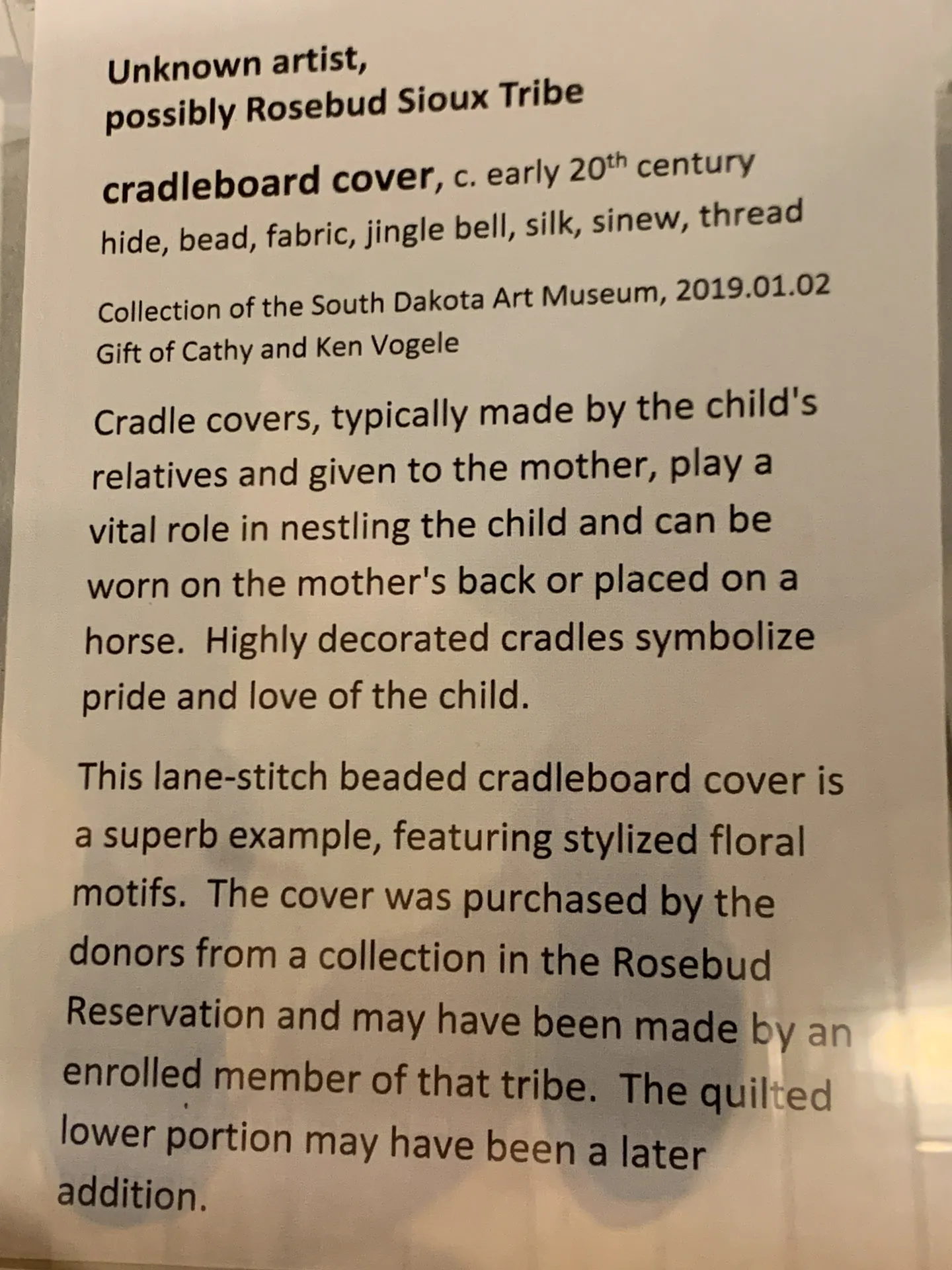 An information plaque about a cradleboard cover. It mentions the piece has hide, beads, jingles, bells, silk, and sinew, dated early 20th century. The text describes its cultural significance and decorative elements, with details about the museum collection and donation.