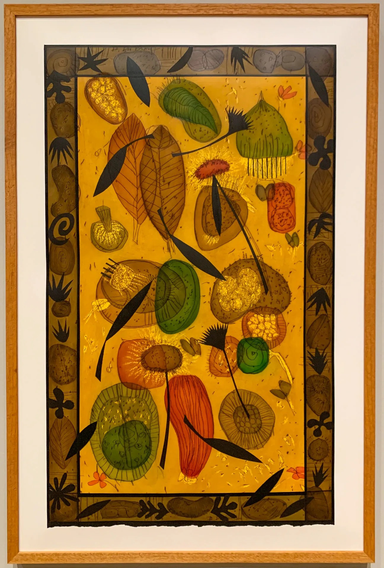 Framed artwork featuring abstract, colorful leaves and shapes on a textured golden background. The border is decorated with black, geometric patterns and symbols. The composition includes green, orange, and brown hues, with intricate details and overlapping elements.