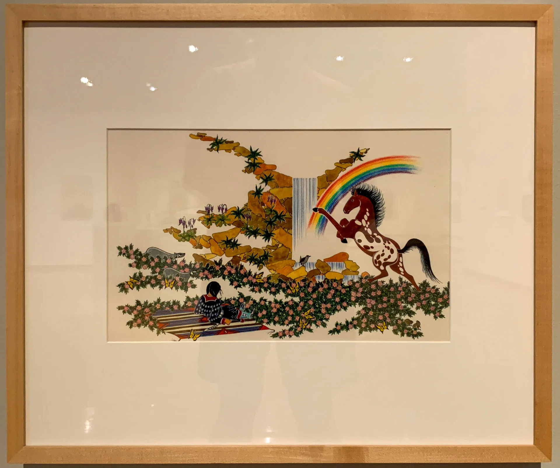 Framed artwork showing a girl reading on a patterned blanket beneath a tree with colorful foliage. A mythical horse with a rainbow mane leaps near a waterfall. Vibrant colors and detailed patterns dominate the scene, framed in white matting with a light wood border.