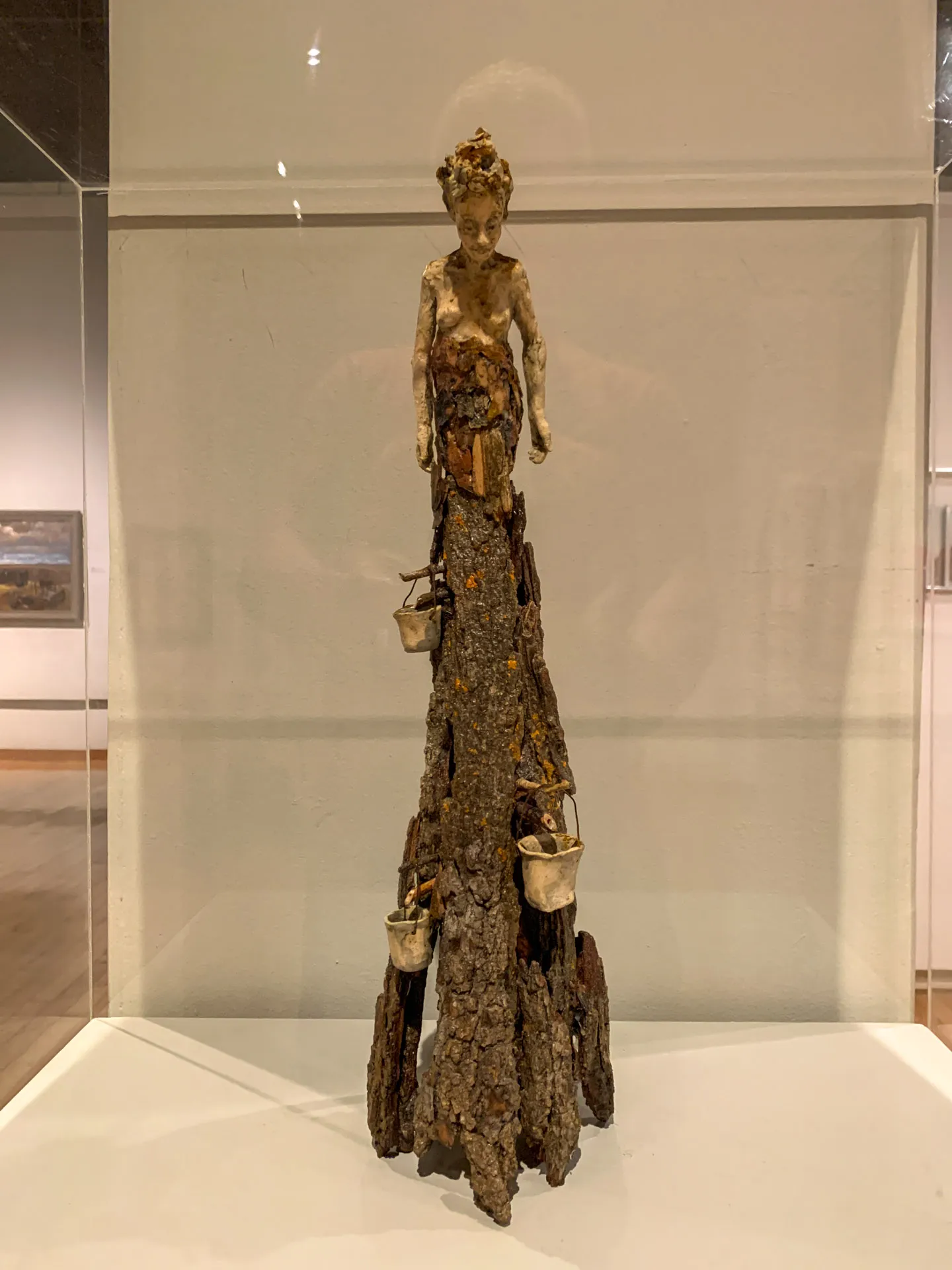 Sculpture of a figure with a weathered, humanoid form standing atop a tall, jagged tree stump. The figure holds small objects in its outstretched hands, with additional objects hanging from the stump. Displayed in a glass case in an art gallery with framed artworks in the background.