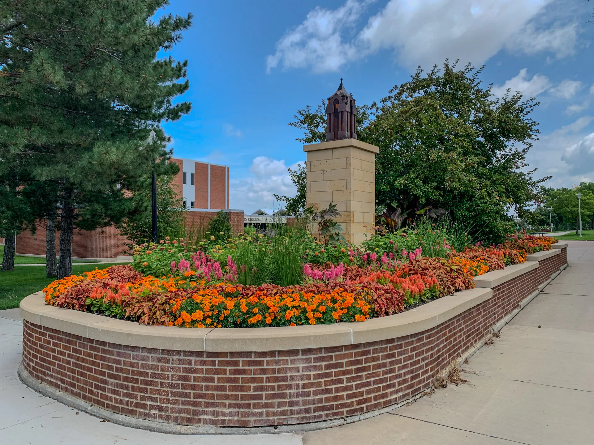 A curved brick planter filled with vibrant orange, pink, and yellow flowers, along with green foliage, surrounds a tall, square stone pedestal topped by a metal sculpture. The scene is set against a backdrop of trees and a red brick building under a partly cloudy sky.