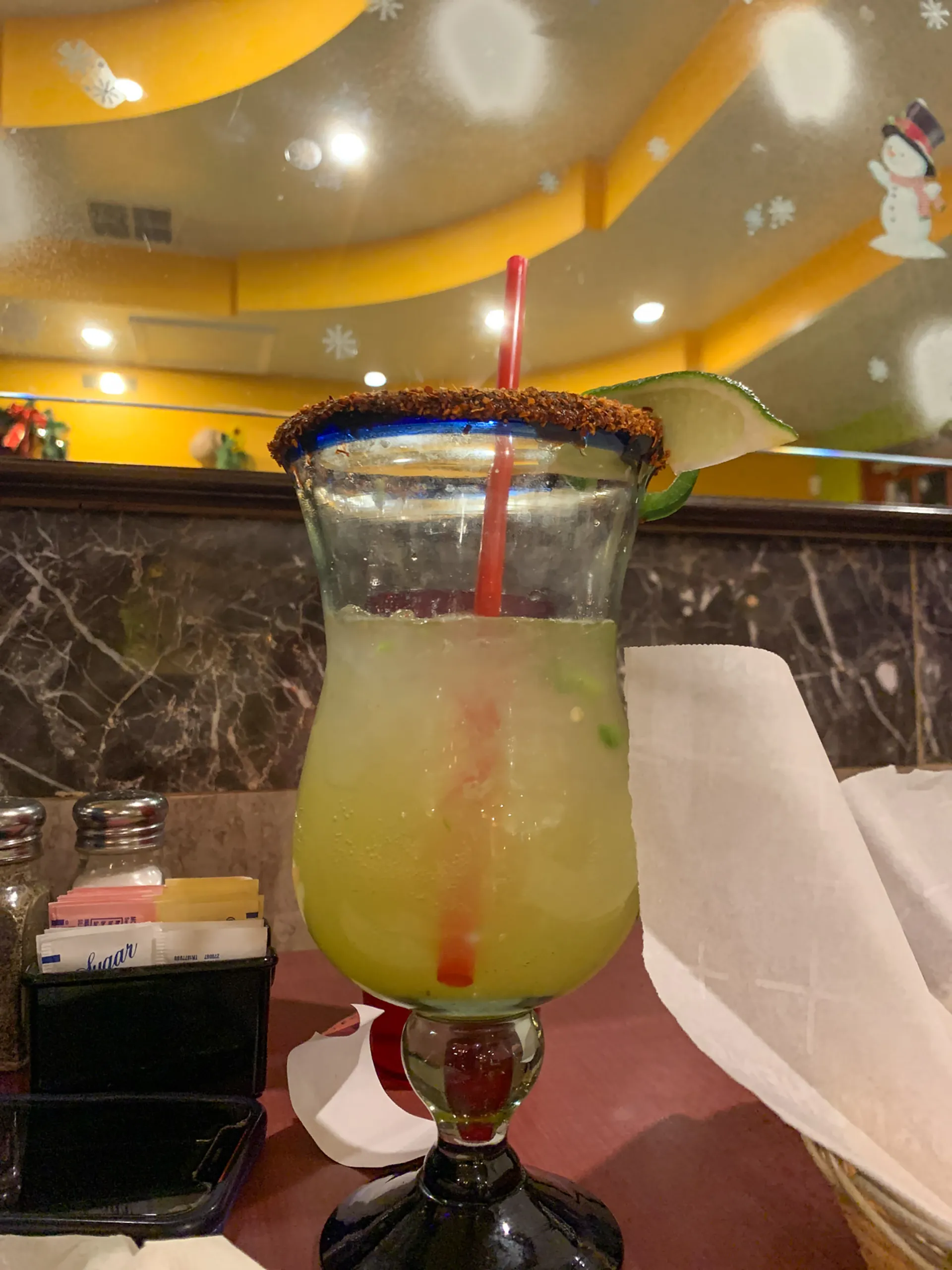 A large margarita glass with a chili powder rim and lime wedge, filled with a light green drink and ice, sits on the table during a Farewell Dinner. A red straw stands out as condiments, a mirror, and yellow ceiling decor complete the restaurant scene.