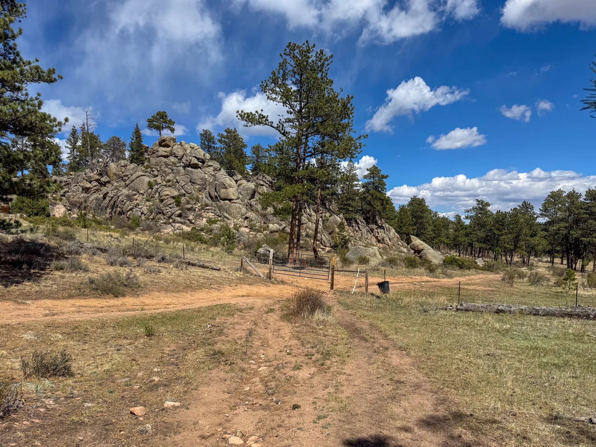 A dirt path forks near a wire fence and gate, inviting hikers toward rocky outcrops and scattered pines beneath white clouds. This serene scene along the Granite Ridge Trail in Roosevelt National Forest captures a tranquil, rural setting.