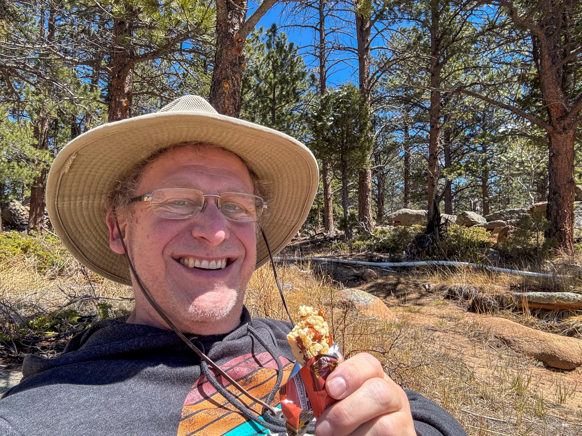 A smiling person wearing glasses, a wide-brimmed hat, and a hoodie holds a granola bar. They are outdoors on the Granite Ridge Trail in Roosevelt National Forest, surrounded by pine trees and rocks, enjoying a sunny day.