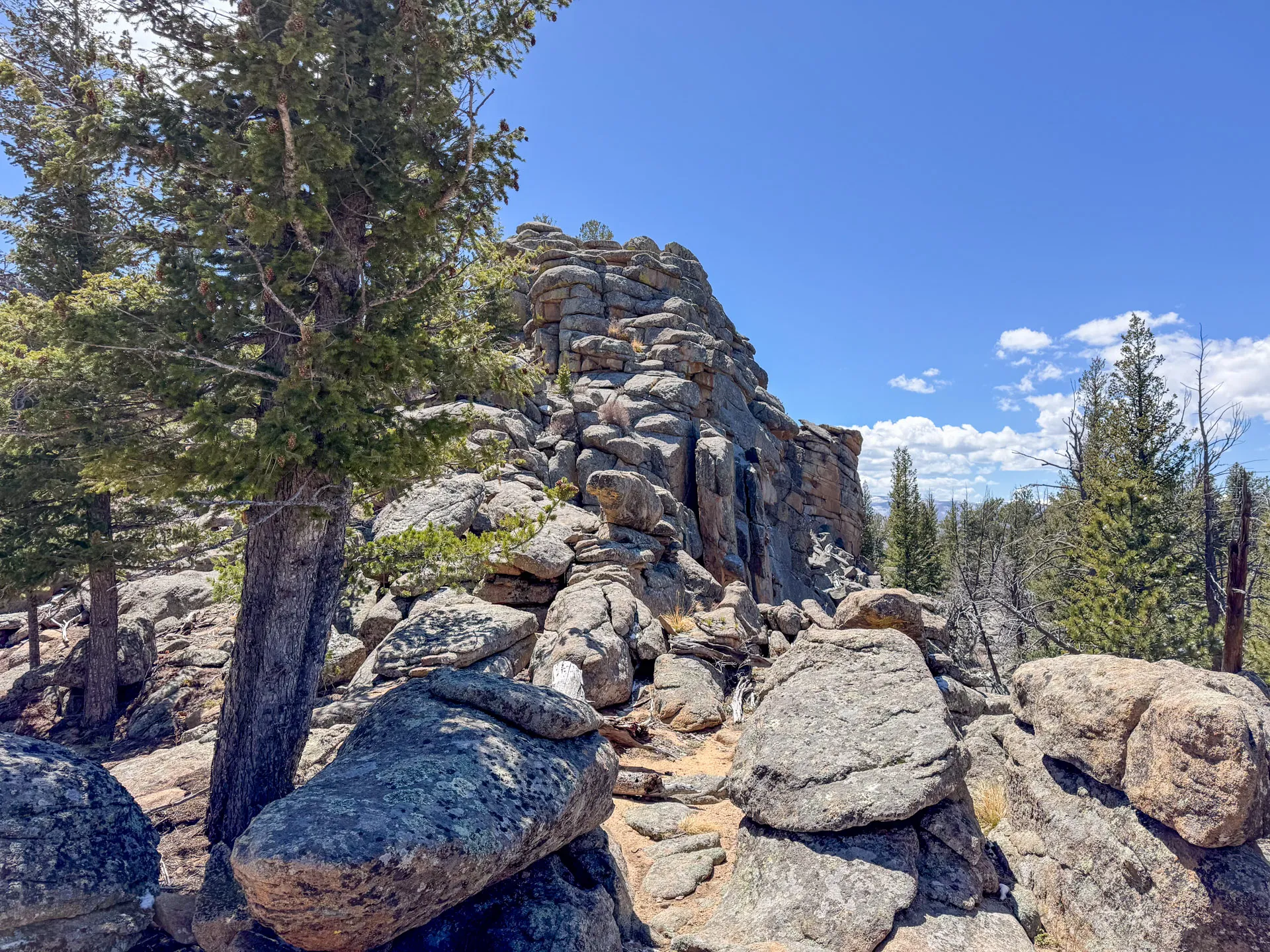 Large rocky outcrop with jagged boulders and rough stones among pine trees under a clear blue sky in Roosevelt National Forest. Sunlight casts shadows on the rocks, with distant clouds visible beyond the Granite Ridge Trail.