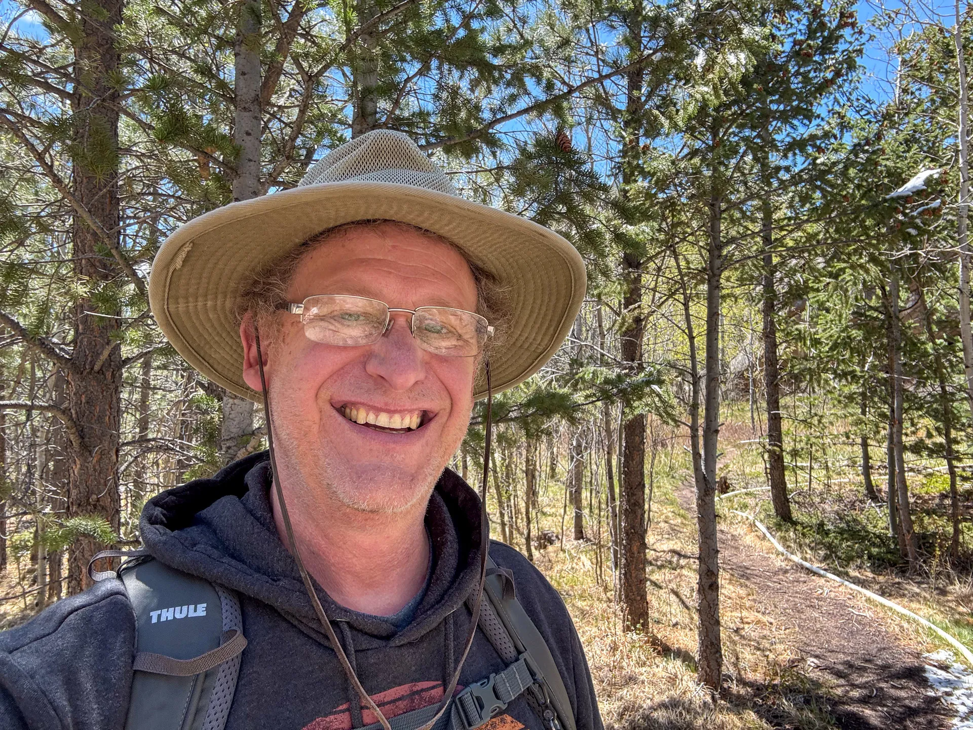 A smiling person wearing glasses, a wide-brimmed hat, and a backpack stands on the Granite Ridge Trail in Roosevelt National Forest, surrounded by tall pine trees and sunlight, enjoying a hike on a clear day.