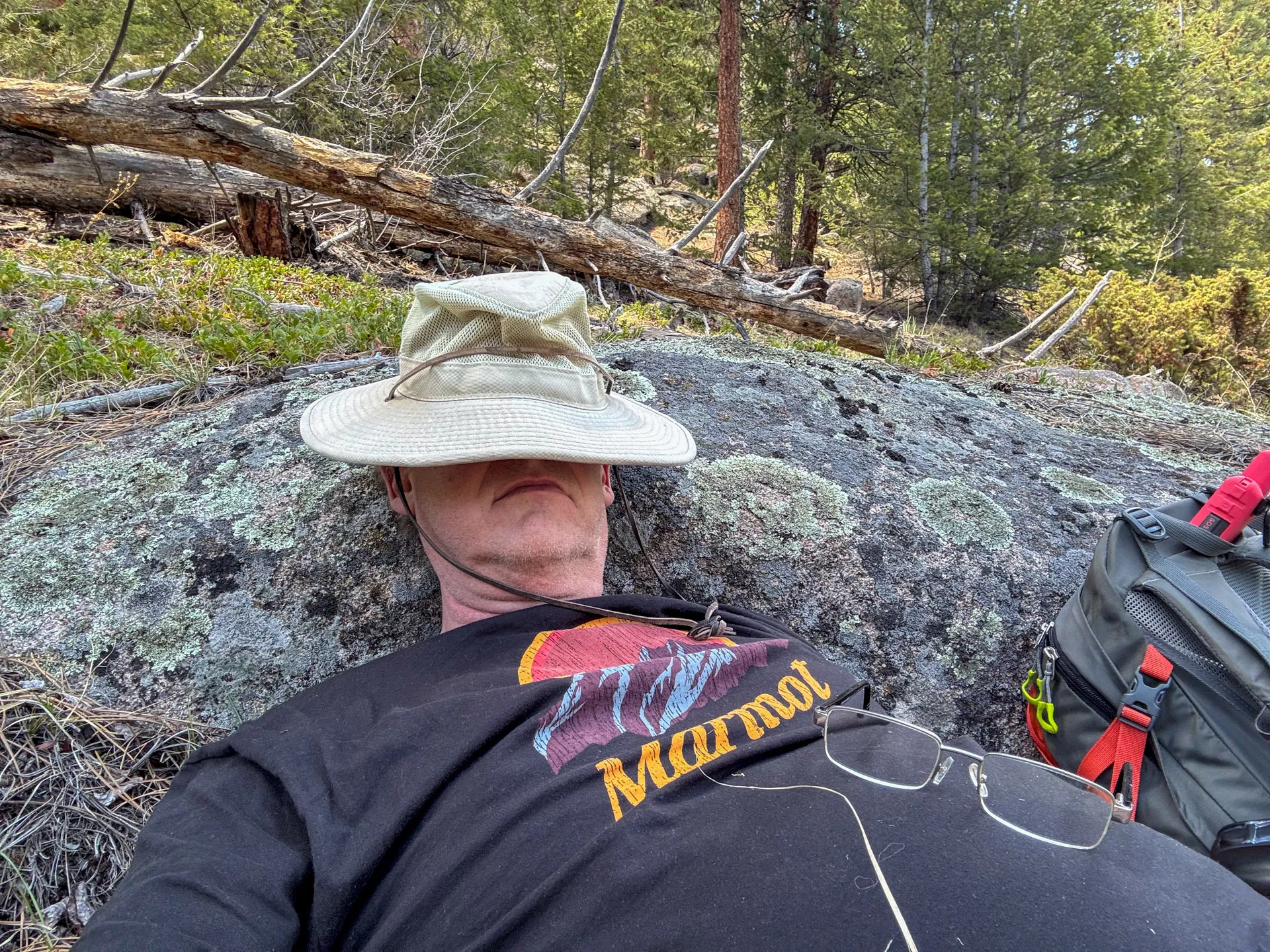 A person wearing a wide-brimmed hat and a black "Marmot" t-shirt lies on a large rock in a forest, relaxing after hiking the Frog Pond Trail. Their face is shaded, glasses rest on their chest, and a backpack sits nearby among grass and fallen branches.