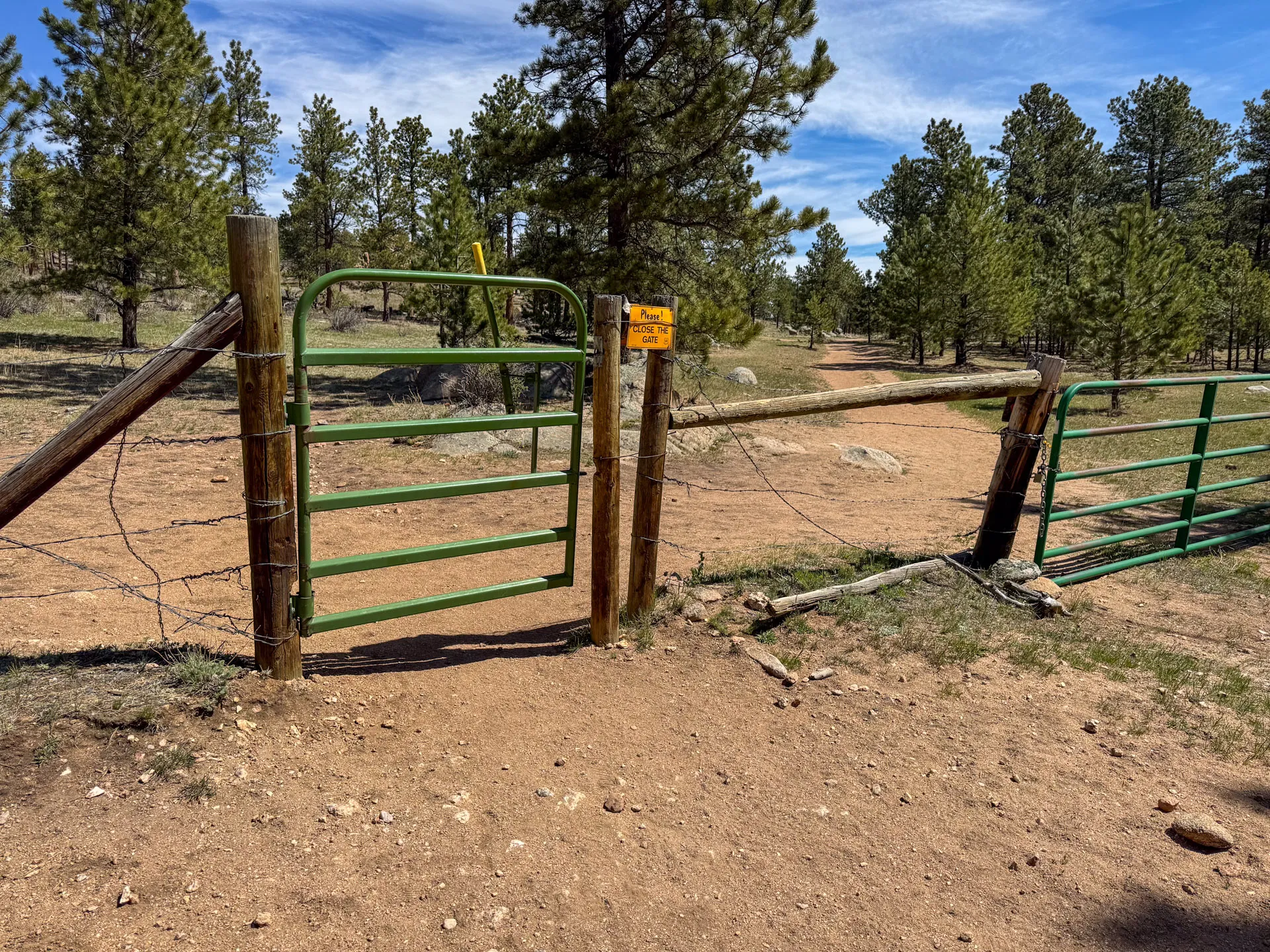 A green metal gate marks the dirt path near in a pine forest, with a yellow "Hiker Access" sign on a wooden post. Another gate stands partially open; rocky ground and trees stretch beneath a blue sky streaked with wispy clouds.