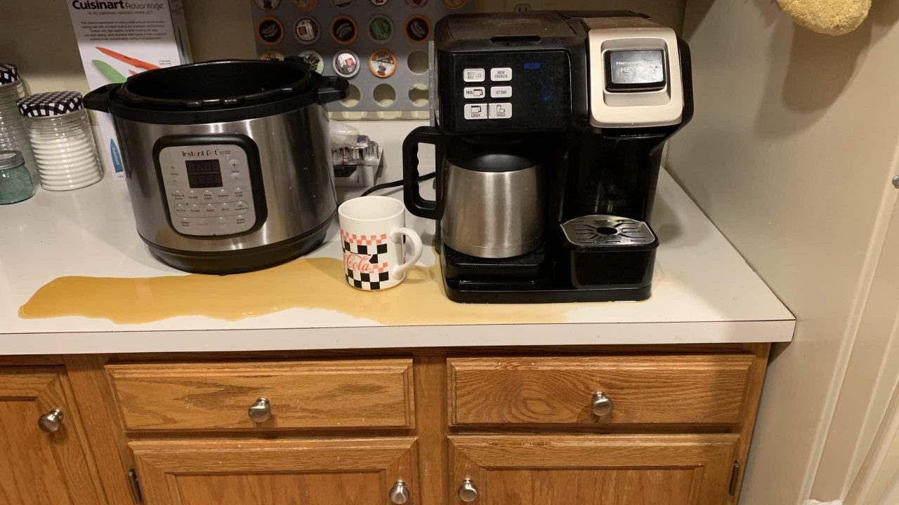 Spilled coffee on a white kitchen countertop with a black and silver coffee maker and a silver multi-cooker sits beside a mug near the edge. With wooden cabinets below, the decision made to install a wall-mounted pod holder with various coffee pods adds functionality and style.
