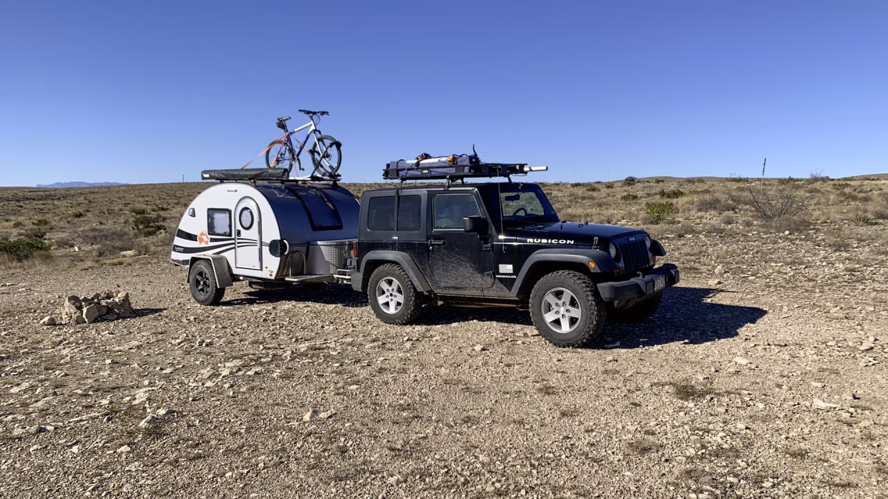 A black Jeep Rubicon with a teardrop trailer attached is parked on rocky BLM Land under a clear blue sky near Carlsbad. Two bicycles are mounted on the trailer's roof, with sparse desert vegetation and distant hills in the background.