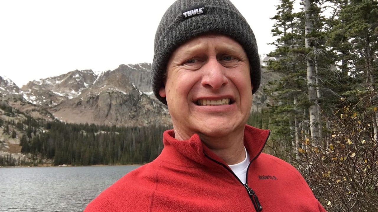 A person wearing a red jacket and gray beanie is smiling awkwardly, as if faced with a 404 error. They stand in front of a scenic mountain lake, where snow-capped peaks and dense evergreen trees create a picturesque background under an overcast sky.
