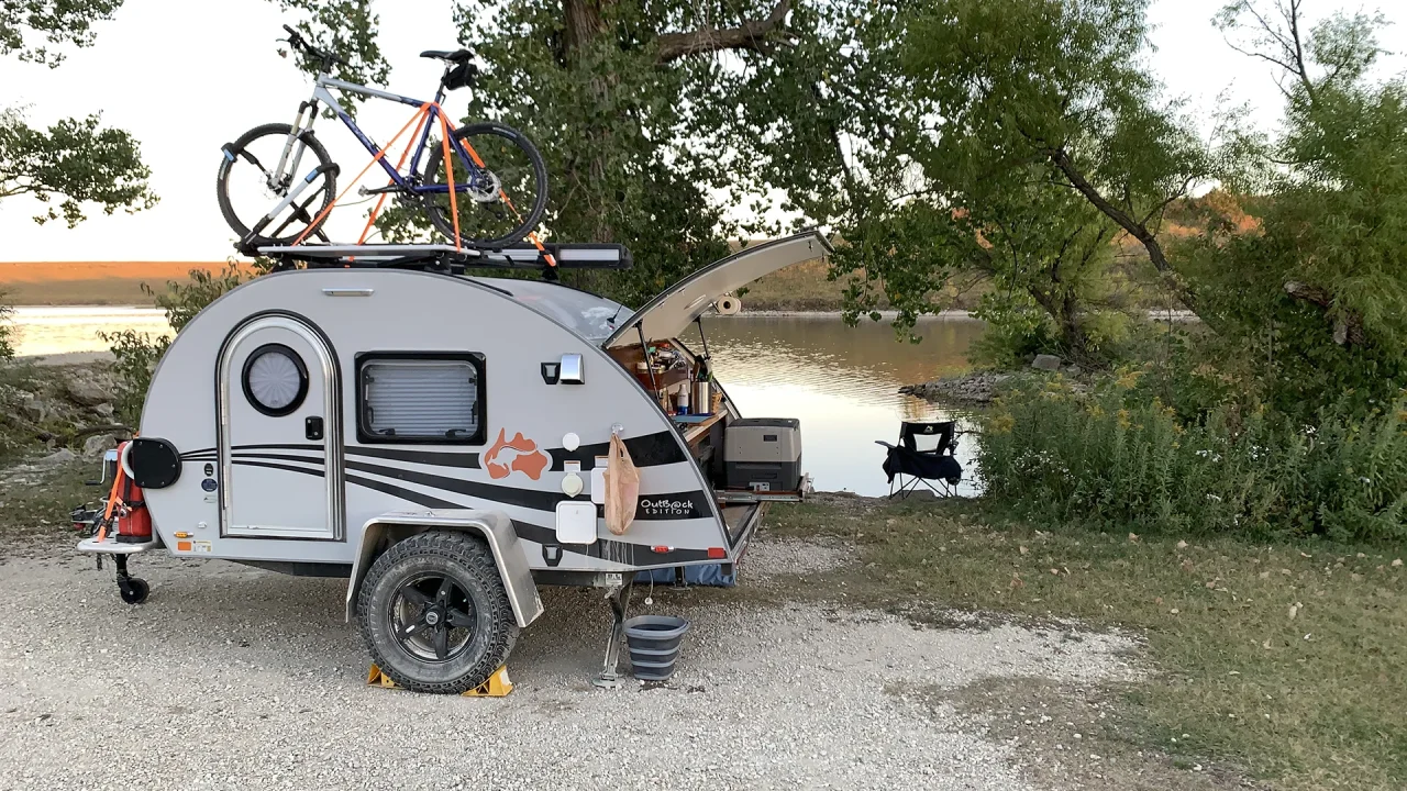 A small travel trailer is parked by a lake with a bicycle mounted on its roof. The trailer's hatch is open, revealing a kitchen area. An outdoor chair and a table are set up beside the lake.