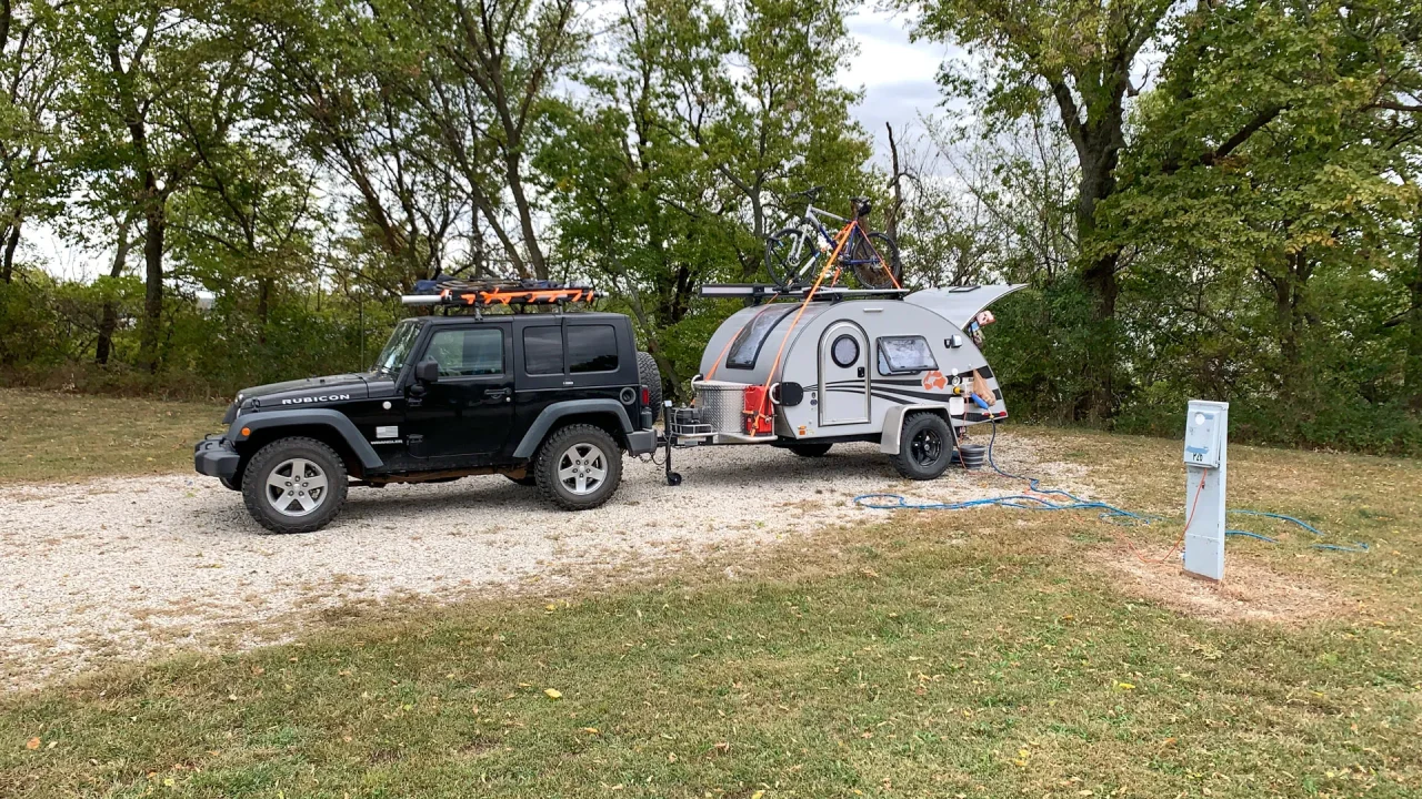 A black Jeep is parked on a gravel path at Clinton State Park, towing a teardrop trailer. The trailer is equipped with a rack holding gear. The scene unfolds in a grassy area with tall, green trees as the backdrop, and an electrical hookup station is visible nearby.