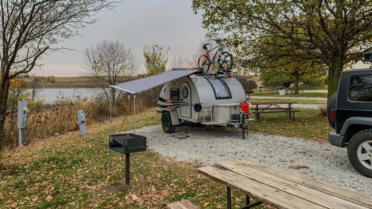 A teardrop camper with an awning is nestled at a Czechland Lake campsite, surrounded by trees adorned with autumn leaves. Bicycles are mounted on its roof, while a picnic table and grill invite outdoor meals. A vehicle rests beside the camper under an overcast sky, completing the serene lakeside scene.
