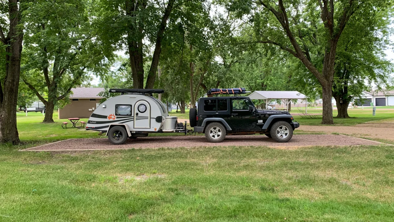 A black jeep with a small white camper trailer is parked in a grassy area under tall green trees at Freeman City Park, near picnic tables and buildings in the background.