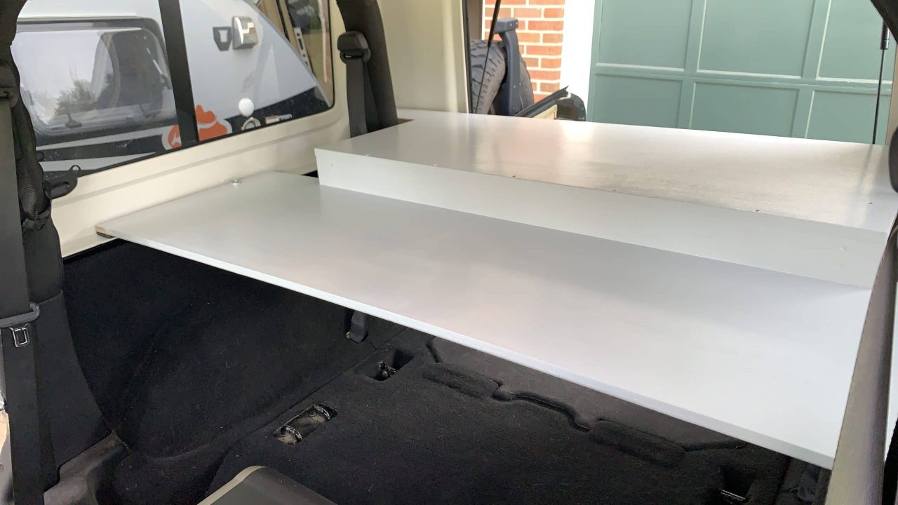 The image shows the rear interior of a vehicle, with a flat, retractable shelf or cargo platform installed. The shelf extends from the backseat area, creating additional storage space above the vehicle's floor. A garage door and part of another vehicle are visible in the background.