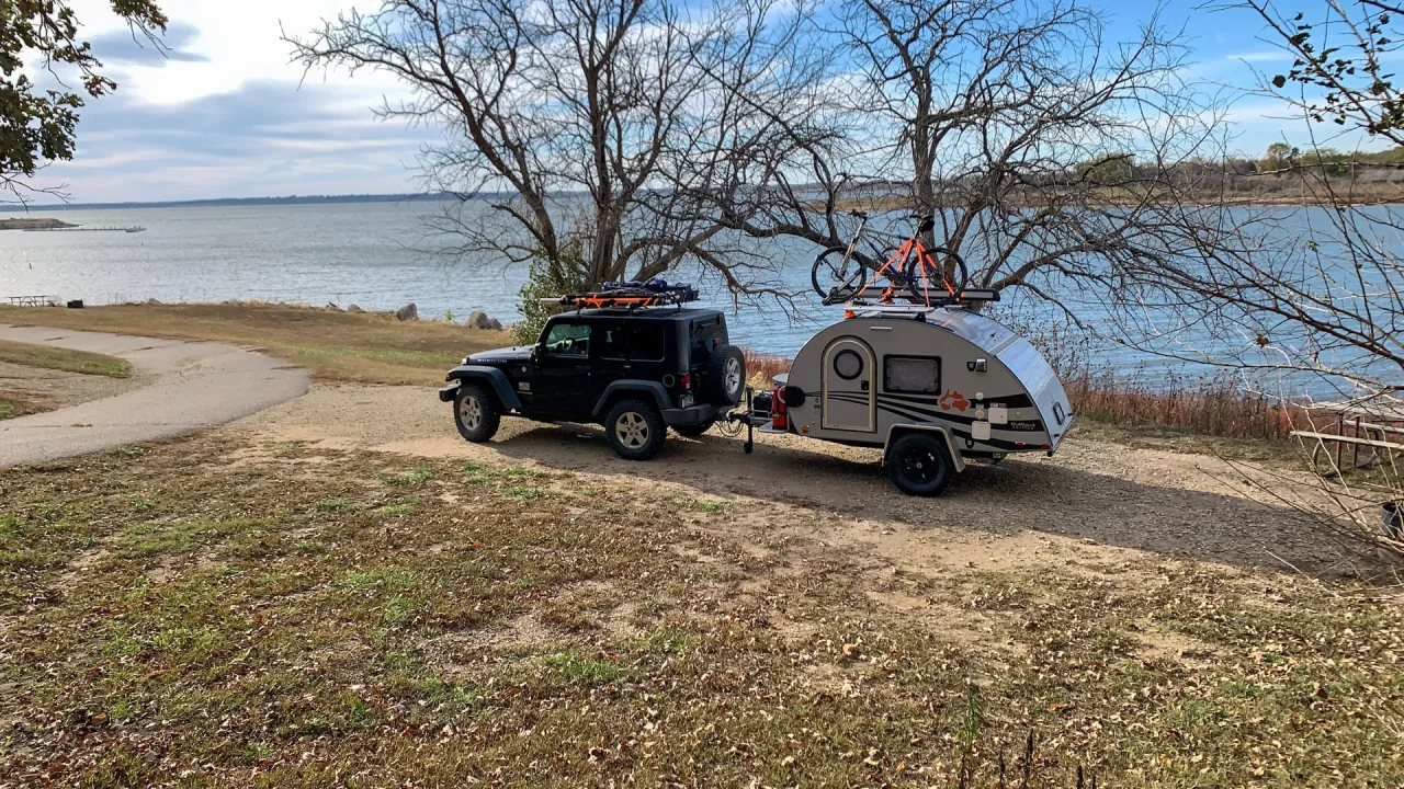 A black Jeep with an attached teardrop camper is parked by the calm lakeside of Milford State Park. Two bicycles are mounted on the camper's roof, surrounded by bare trees and a grassy, slightly brown landscape, suggesting late autumn. The mostly clear sky and a few clouds reflect in the tranquil water.