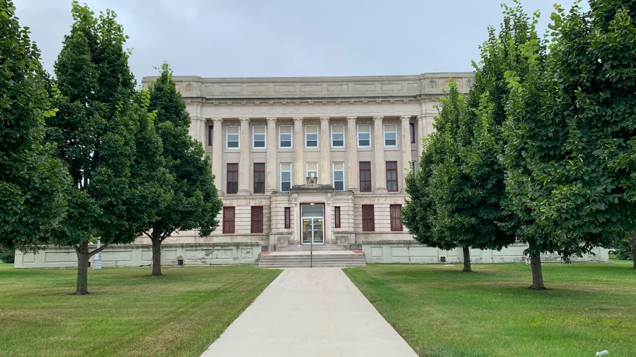 A historic, beige stone courthouse with six columns stands at the end of a concrete walkway in Flandreau, SD. Flanked by manicured green lawns and tall, leafy trees, the Moody County Courthouse is set under an overcast sky that casts a soft light on its large windows with reddish frames.