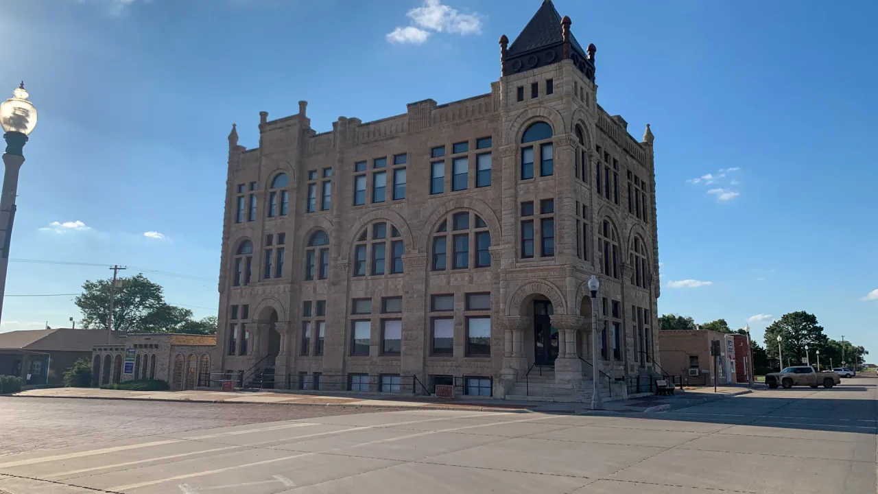 A historic stone building with arched windows and a tower stands at a corner intersection under a clear blue sky.