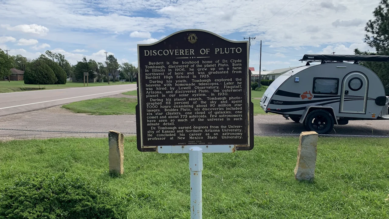 A historical marker titled "Discoverer of Pluto" stands proudly on the grassy roadside. Behind it, a small teardrop trailer waits, hitched to a vehicle under the partly cloudy sky. Trees gently line the road, bearing witness to Clyde Tombaugh's monumental discovery of Pluto.