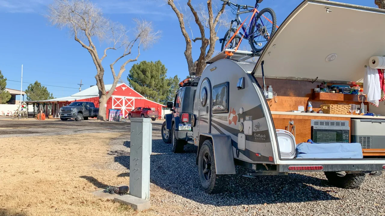 A small teardrop camper is parked on gravel near a grassy area at Red Barn RV Park, its back hatch open to reveal a compact kitchen. A bicycle rests on the roof, with a red barn, leafless trees, and a car nearby under the blue sky.