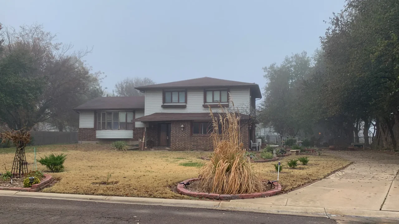 A two-story house with a brick facade and white siding sits on a foggy day in Rose Hill, Kansas. The house evokes a feeling of old charm, with its large, neatly kept lawn and circular driveway. Trees surround the property, their branches slightly visible through the misty, overcast sky.