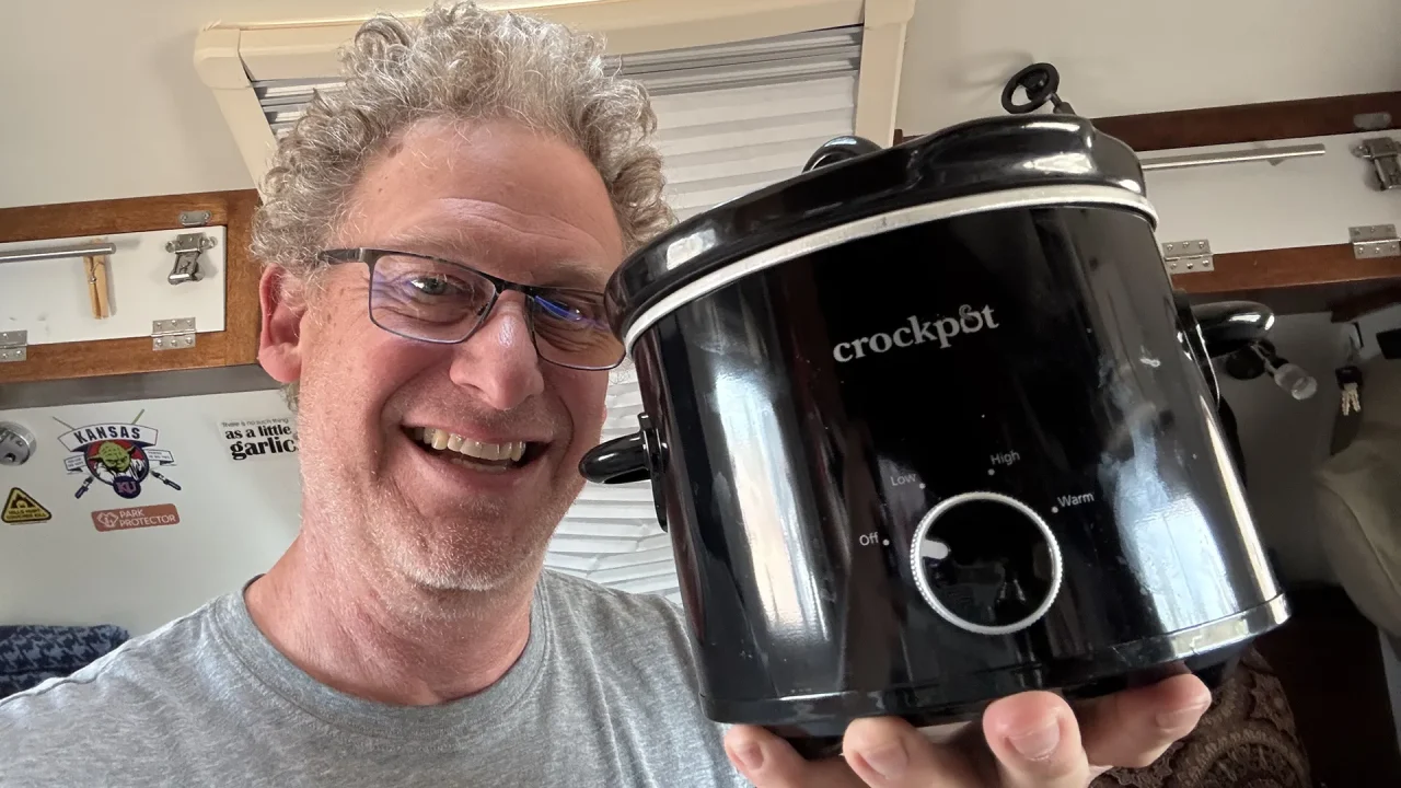 A person with curly hair and glasses grins while holding a black Crockpot, dreaming of making exotic dishes like Pineapple-infused recipes. They are indoors, with a wall of stickers behind them, including a garlic design. The Crockpot's control knob offers settings for warm, low, and high.
