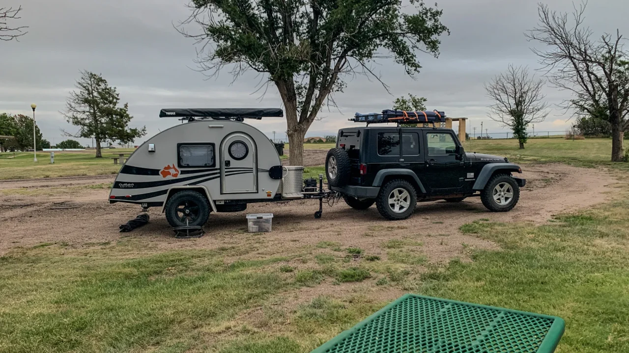 A black Jeep is parked on a dirt area towing a small, white teardrop camper in St. Francis City Park. Nearby trees and a picnic table are visible under the cloudy sky.