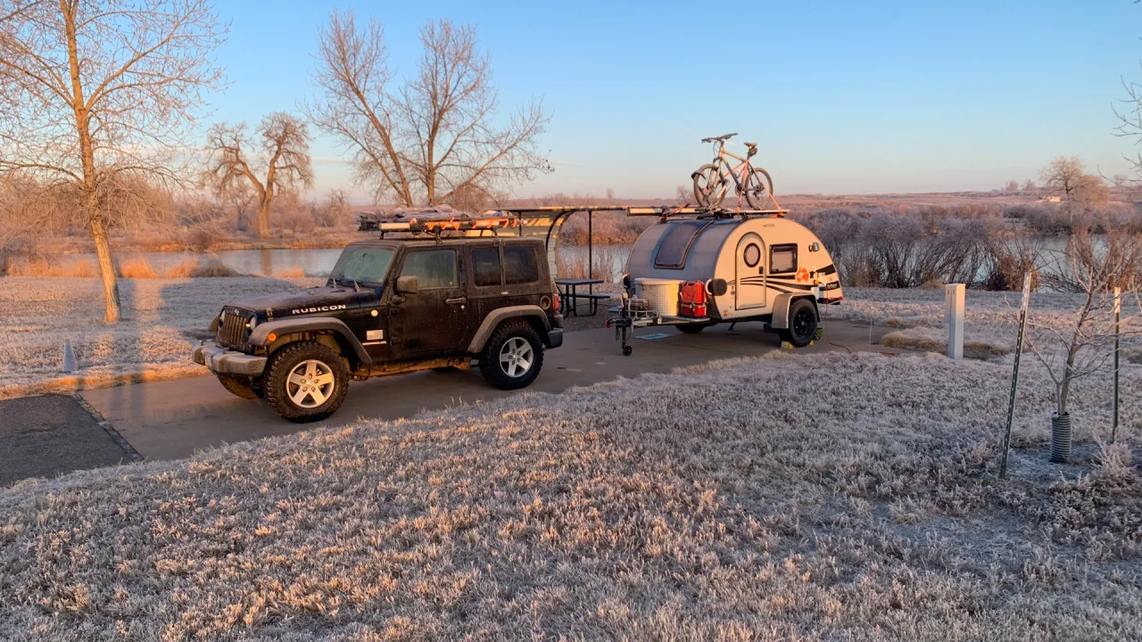 A black Jeep with a roof rack is parked on a frosty driveway at St Vrain State Park, towing a small teardrop trailer with a bike mounted on top. Bare trees and frosty grass glisten in the golden sunrise under a clear blue sky.
