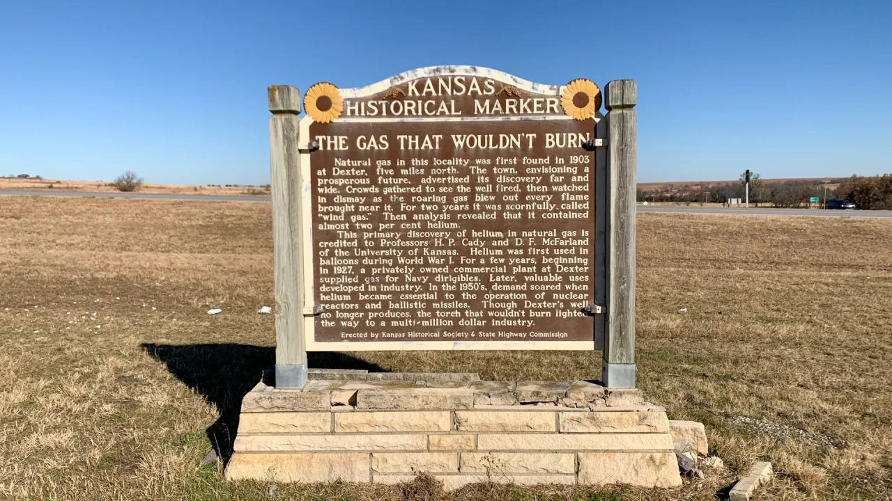 A Kansas Historical Marker stands on a grassy area under a clear blue sky. The sign, adorned with sunflower emblems, tells the tale of “The Gas That Wouldn’t Burn,” highlighting an 1890s natural gas discovery in Dexter, Kansas. A road and open fields unfold gracefully in the background.