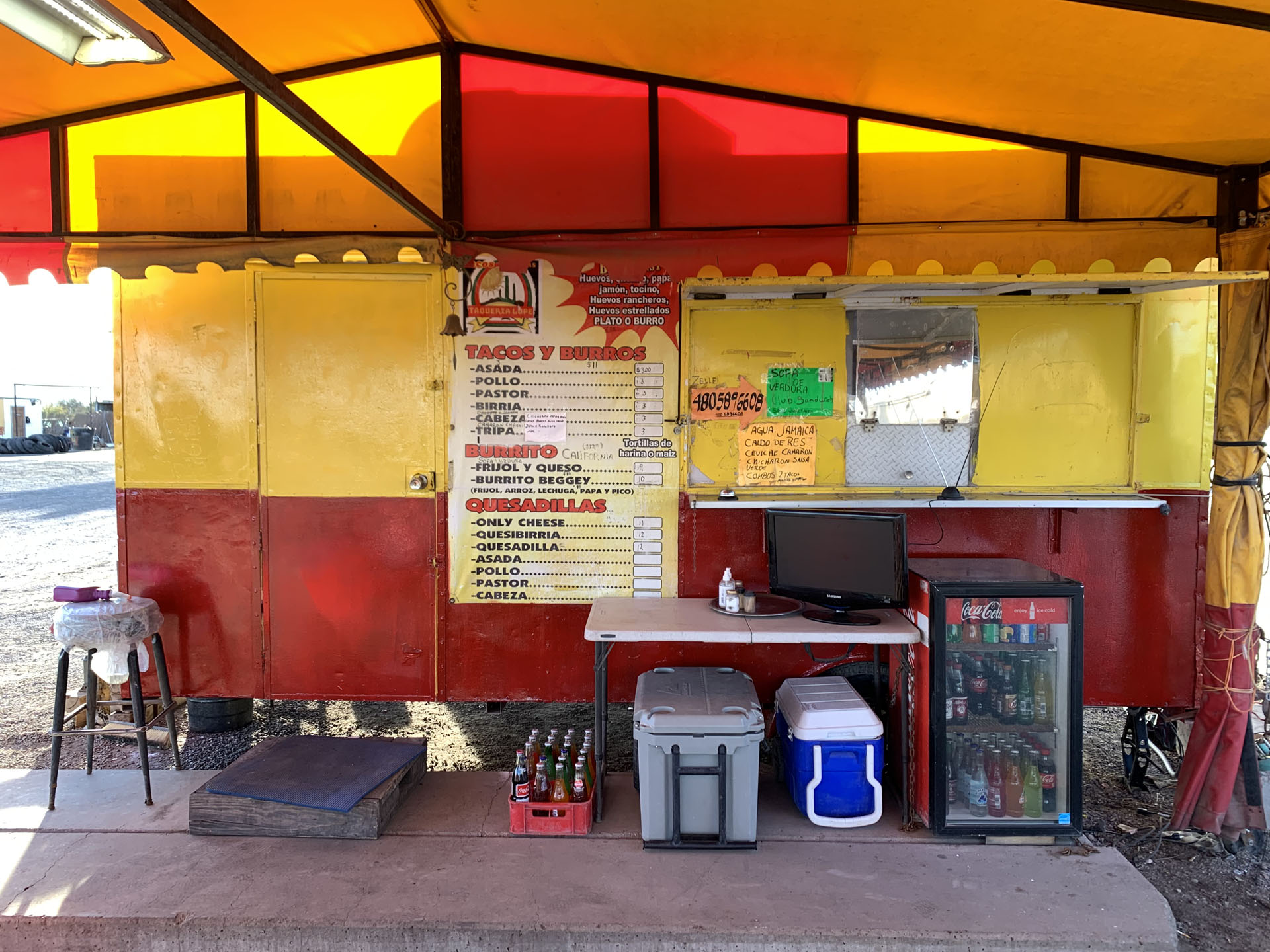 Lupe’s Taqueria is a small outdoor taco stand painted red and yellow beneath a canopy. Beside the serving window, handwritten signs and a menu are posted. In front, there’s a table with a TV, cooler, soda bottles, and a mini fridge filled with drinks.