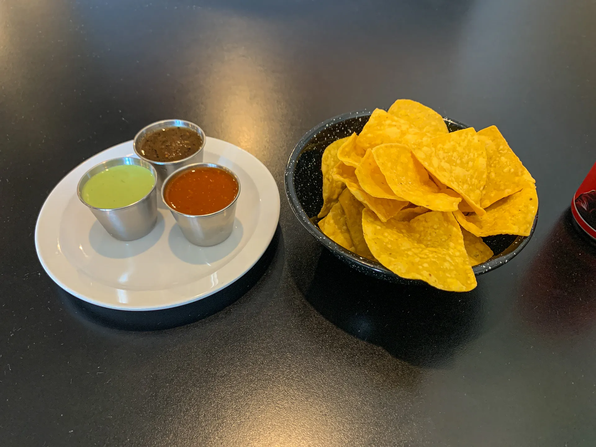 A basket of yellow tortilla chips beside a plate holding three metal cups containing green, brown, and red salsa on a black surface.