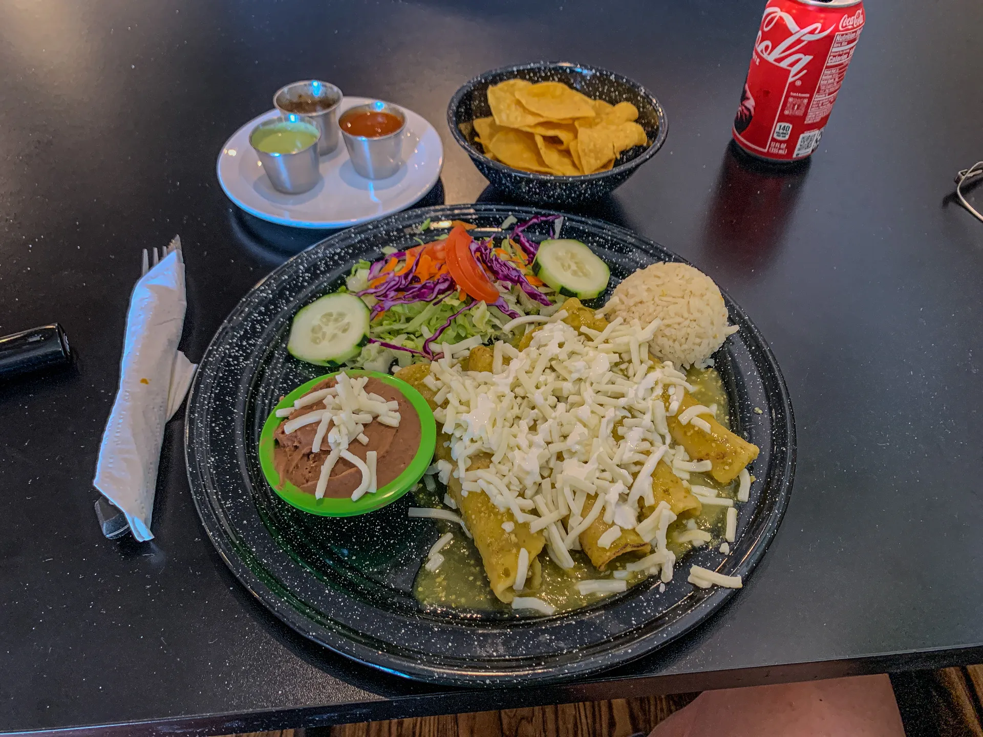 A plate of enchiladas topped with cheese, served with refried beans, rice, salad, and cucumber slices, accompanied by a smaller plate with dipping sauces, tortilla chips, and a can of Coca-Cola.