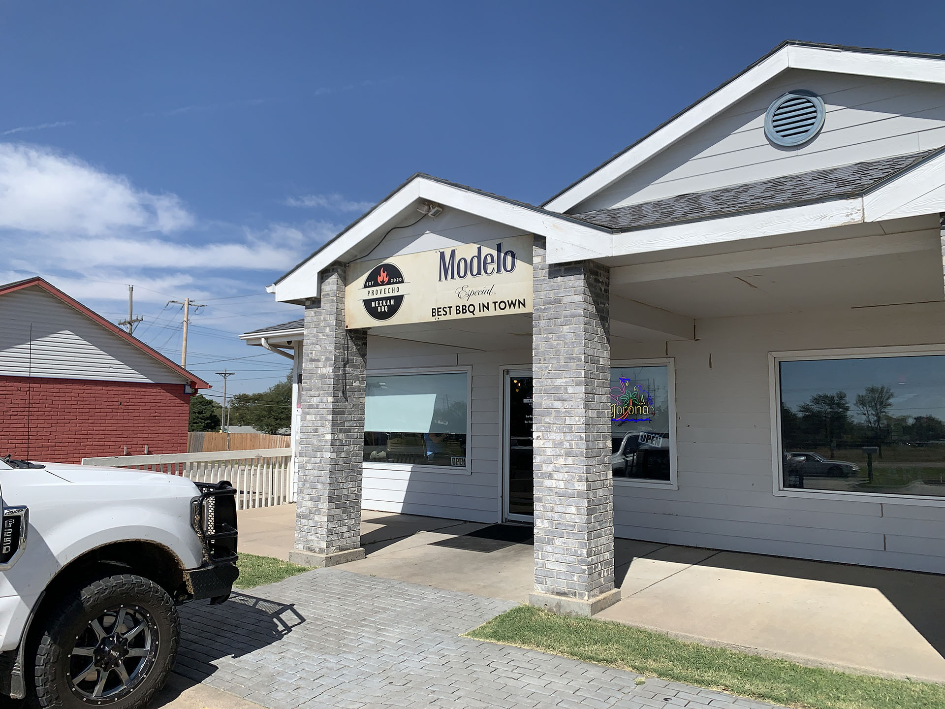 A white building with a sign advertising "Modelo - Best BBQ in Town" above the entrance, a truck parked in front, and a clear blue sky in the background.