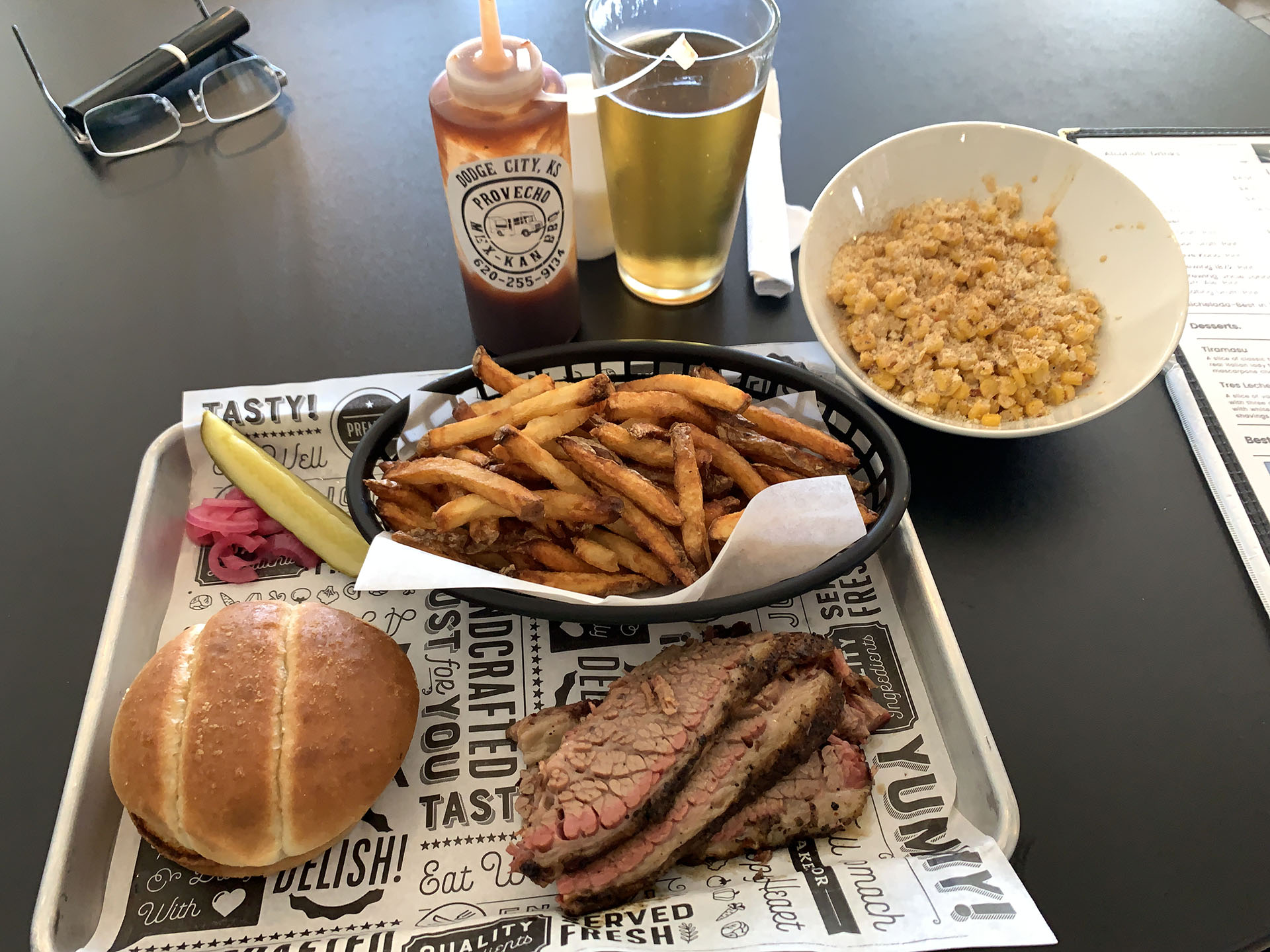 A meal consisting of sliced brisket, French fries, a bun, a pickle spear, corn casserole, a bottle of barbecue sauce, and a glass of beer is served on a tray. Glasses and a menu are set on the table.