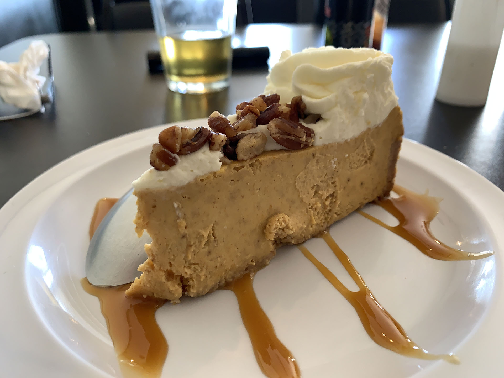 A slice of pumpkin cheesecake topped with whipped cream and pecans, drizzled with caramel sauce, on a white plate. A glass of drink in the background.