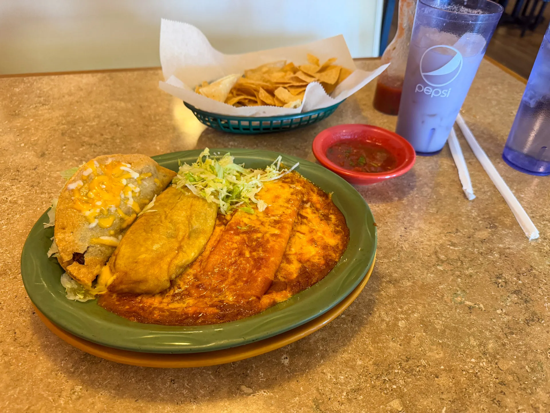 A plate with an enchilada, taco, and chile relleno topped with cheese and shredded lettuce on a green dish. A side of crispy tortilla chips in a basket and a cup of Pepsi are on the table. A smaller cup with straws is next to the drink. Salsa is served in a small dish.