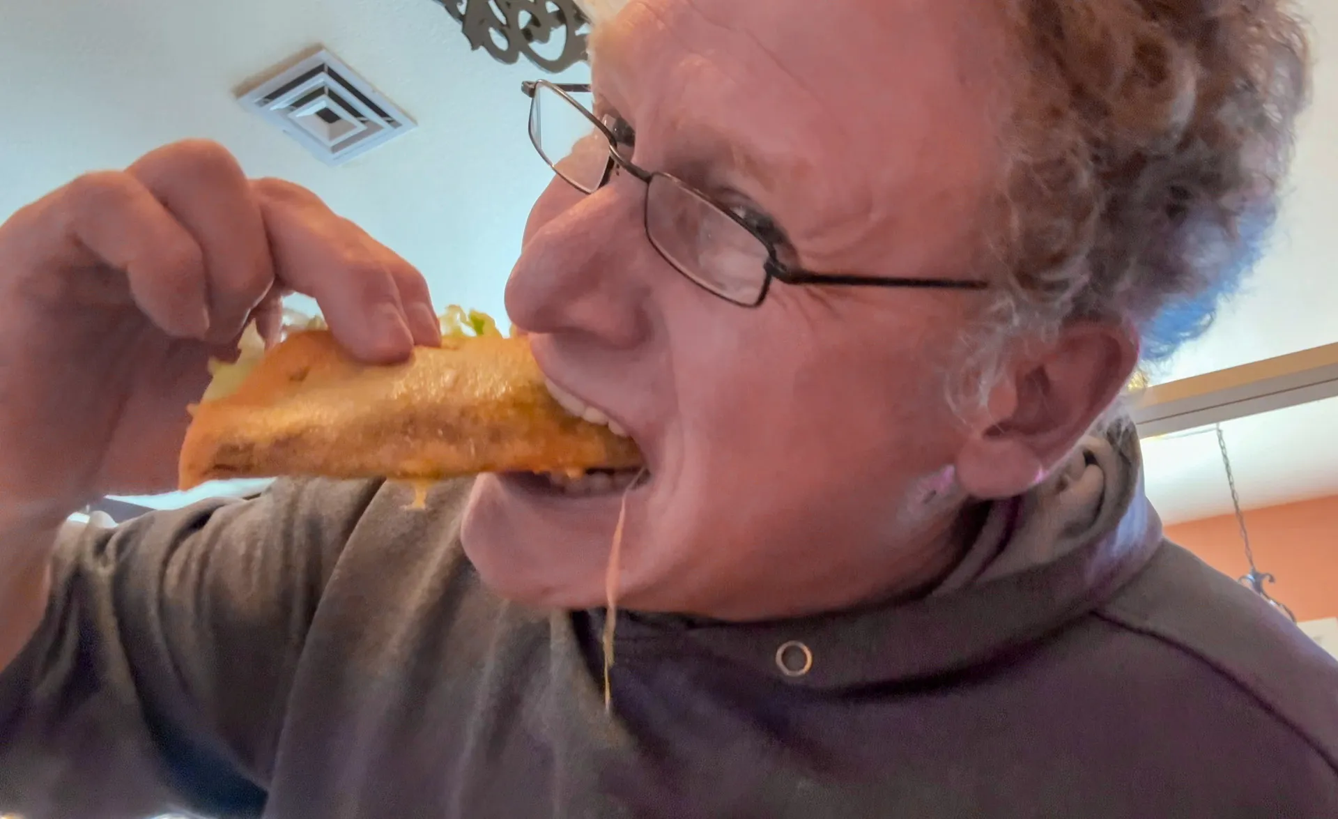 Man wearing glasses and a hoodie leans in, biting into a taco. Melted cheese stretches from the taco, emphasizing its cheesy filling. He appears to be enjoying the food, which has a crisp shell. The background shows a ceiling vent and decorative wall piece indoors.