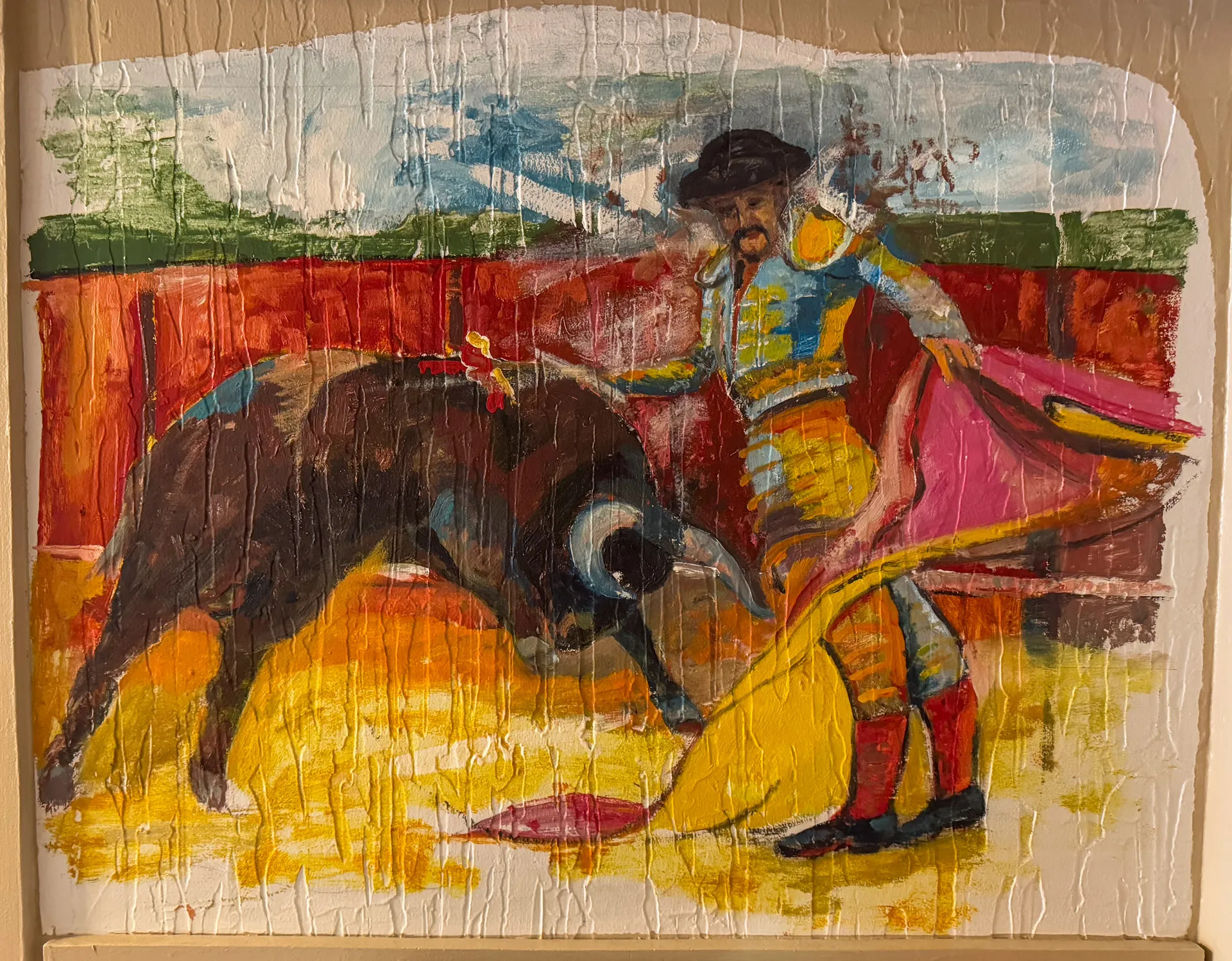 A colorful painting depicts a bullfight. The matador, in a traditional sequined costume, holds a pink and yellow cape, facing a charging bull. The arena is surrounded by a red barrier with a cloudy sky above, creating an intense and dynamic scene.